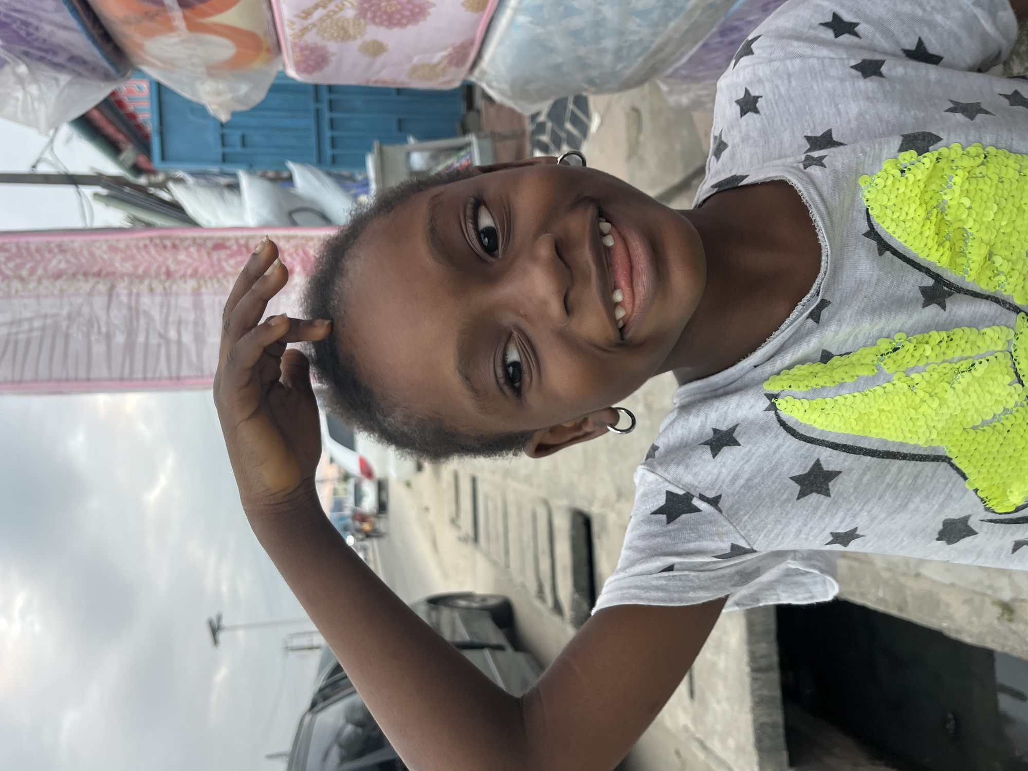 In the city of Ado, Nigeria, a young Black girl with hoop earrings is captured in a close-up shot, her face turned to the side with a wide, toothy smile. She is wearing a grey t-shirt adorned with black stars and a bright neon-green sequined design. Her left arm is raised, with her fingers playfully touching the top of her head. The background suggests an urban outdoor environment with cars and a street visible to her left, and stacks of mattresses and a blue gate to her right. The lighting indicates an overcast day.