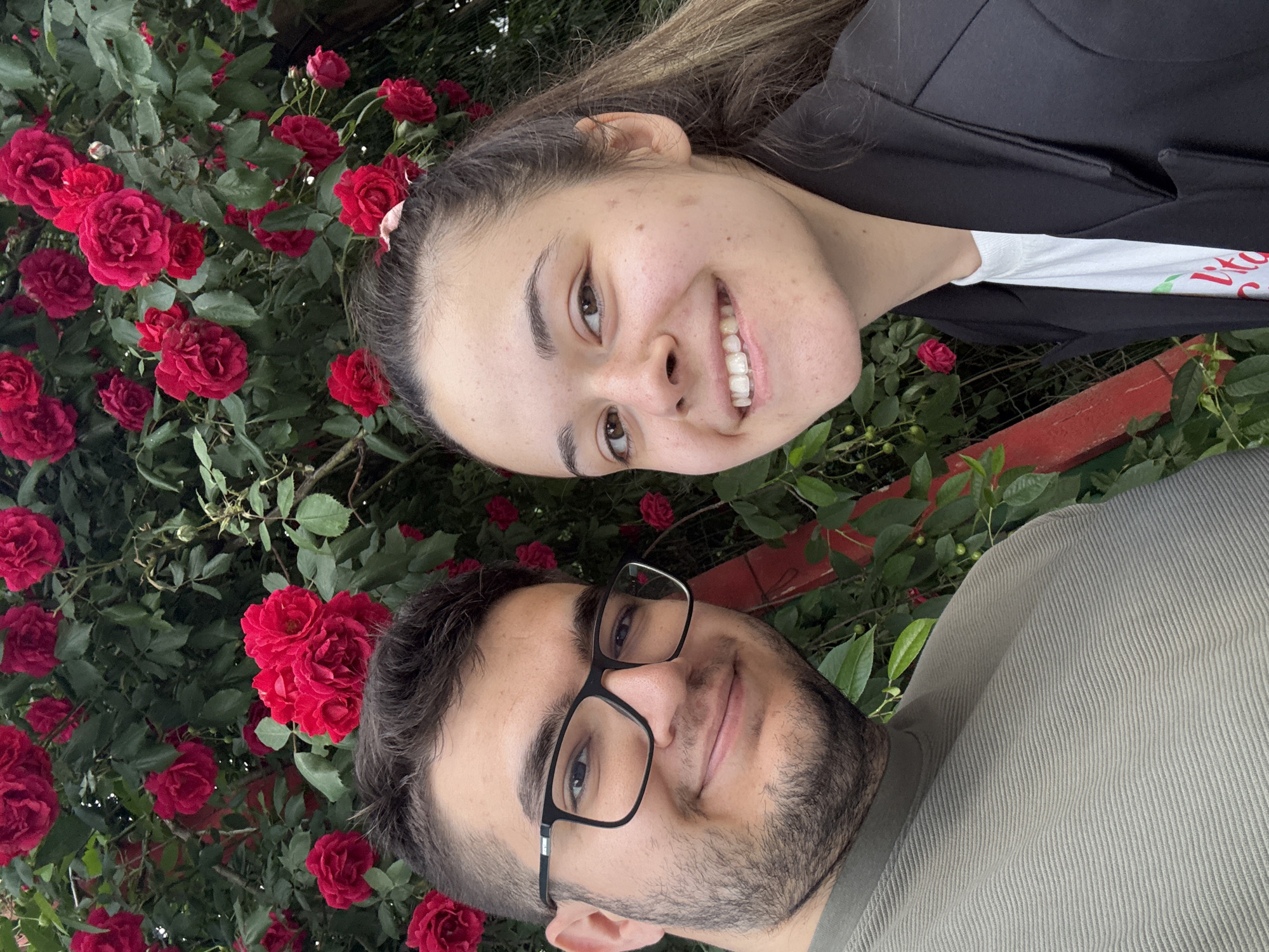 The image, originally captured in a landscape orientation but presented vertically, features a smiling couple positioned outdoors against a backdrop of vibrant red roses and green foliage. The scene appears to be a garden or park in Sofia, Bulgaria.
In the foreground, to the right (if rotated correctly), is a man with short dark hair, a neatly trimmed beard, and black-framed glasses. He has a warm, gentle smile and is looking directly at the camera. He is wearing a light greenish-grey top with a distinct ribbed texture.
Above him and to his left, a woman with long, light brown hair, a bright, wide smile, and natural-looking skin also looks directly at the camera. She wears a dark jacket over a white t-shirt. Partially visible red text on her t-shirt shows "Lit...", possibly part of a word like "Live" or "Life". A small, pink floral hair clip is subtly placed in her hair near her forehead.
The background is dominated by a lush rose bush, densely packed with numerous red roses in various stages of bloom, interspersed with rich green leaves. A reddish-brown fence or railing can be seen peeking through the foliage, particularly between the two individuals. The lighting is soft and natural, indicating an outdoor daytime setting. Both individuals convey happiness and contentment through their smiles.