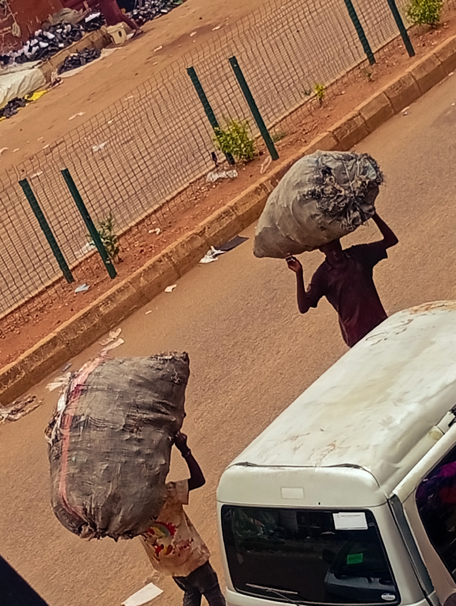 Two individuals are carrying large bags on their heads while walking on a city street. One man is wearing a maroon-colored shirt, while the other is wearing a tan shirt with a rainbow design. A white vehicle is parked on the right side of the frame. A wire fence and sparse vegetation run along the side of the road. The city is identified as Unknown, Unknown.