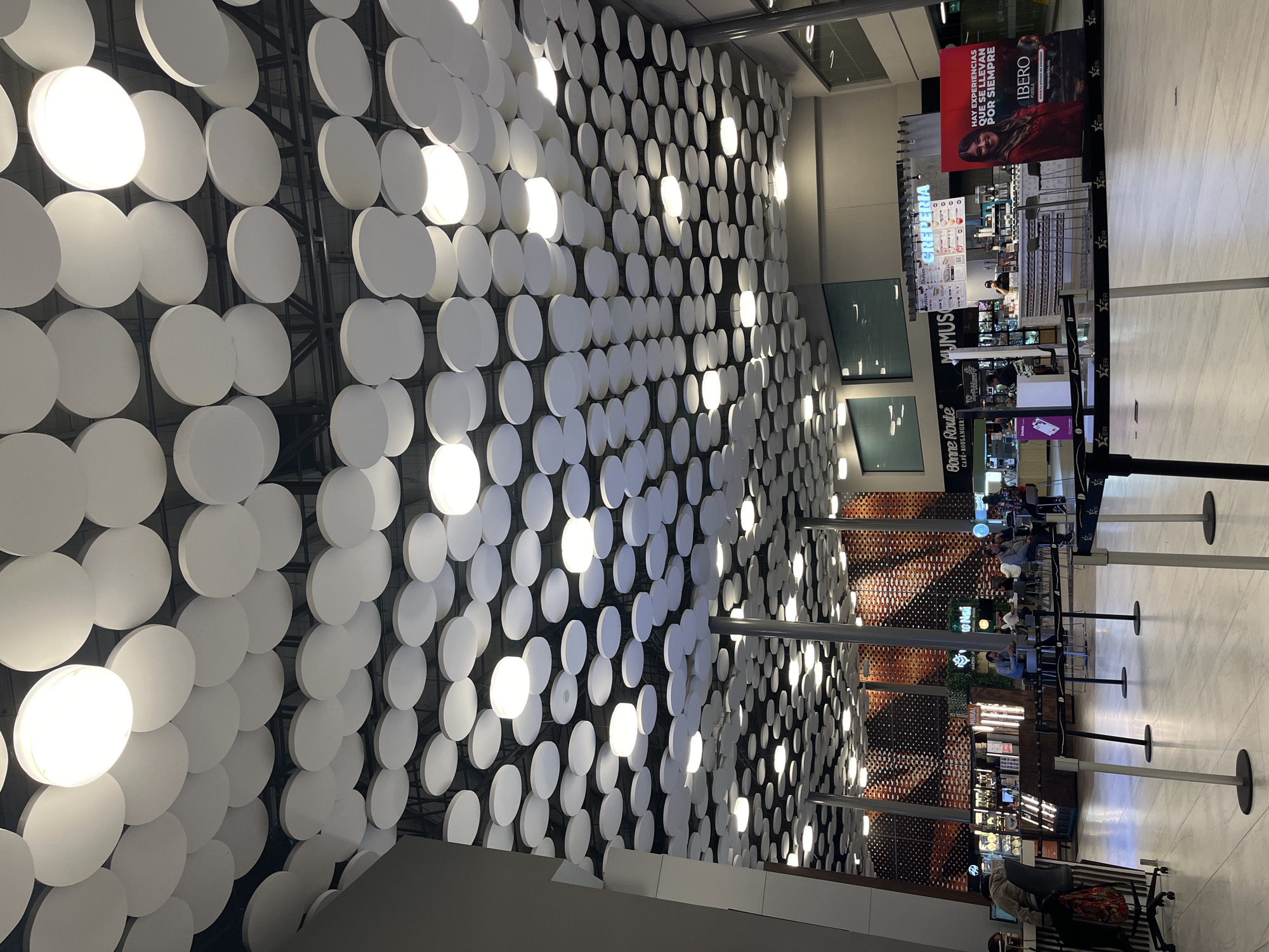 The image depicts the interior of a commercial space, likely an airport or shopping mall, in Puebla, Mexico. A wall covered in numerous illuminated white disks dominates the left side of the frame. To the right, various food and beverage establishments, including "Bonne Route CAFÉ-BOULANGERI", "MUMUS", and "CREPERIA," are visible. An advertisement for "IBERO" Puebla is prominently displayed above one of the establishments. People are seated at tables, and stanchions are set up, possibly indicating waiting areas. The overall scene suggests a modern, well-lit commercial environment.