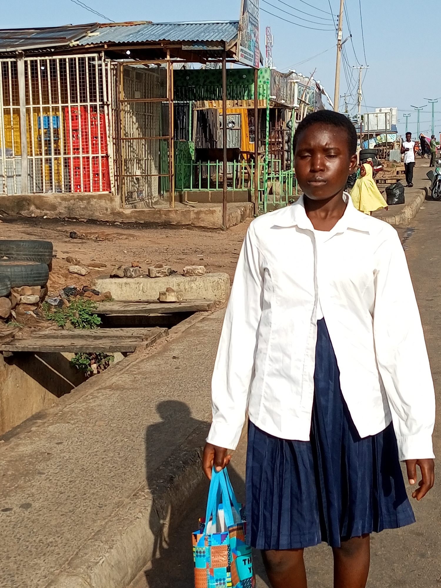 In Garko, Nigeria, a young Black girl is captured in a medium shot, walking on a paved street beside a raised concrete walkway. She is wearing a white button-down shirt over a dark blue pleated skirt and carrying a colorful tote bag with blue straps in her right hand. Her eyes are closed, and her head is slightly tilted downwards. Behind her, the street is lined with market stalls constructed from corrugated metal and wire mesh, displaying various items like red and yellow crates. The ground is uneven and appears to be a mix of dirt and rubble. In the background, other people are visible, along with a motorcycle and telephone poles. The scene is illuminated by natural daylight, casting a shadow of the girl on the walkway.