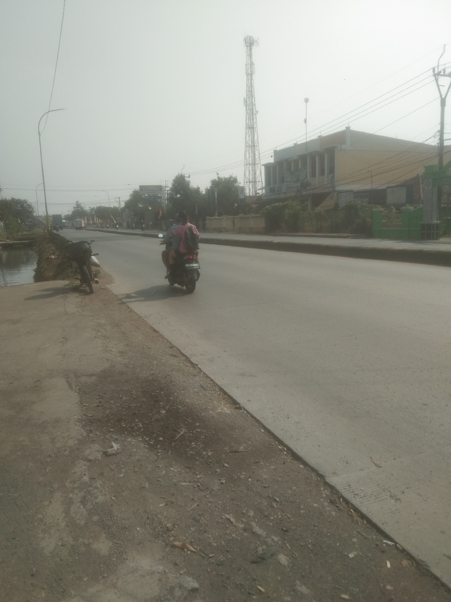 The image shows a roadside scene in Subang, Indonesia, characterized by hazy atmospheric conditions and a wide, paved road. In the foreground, to the left, is a concrete walkway next to a narrow body of water. A parked motorcycle is visible on the edge of the walkway. Further down the road, a person is riding a motorcycle with another passenger on the back. They are traveling away from the camera, moving into the hazy distance. Along the right side of the road, buildings are visible, including a structure with a yellow facade and columns, and a tall cell tower stands prominently against the sky. Trees and smaller buildings line the opposite side of the road, with additional vehicles and utility poles also present. The overall environment suggests a commercial or semi-urban area, with the haze obscuring details and affecting visibility.