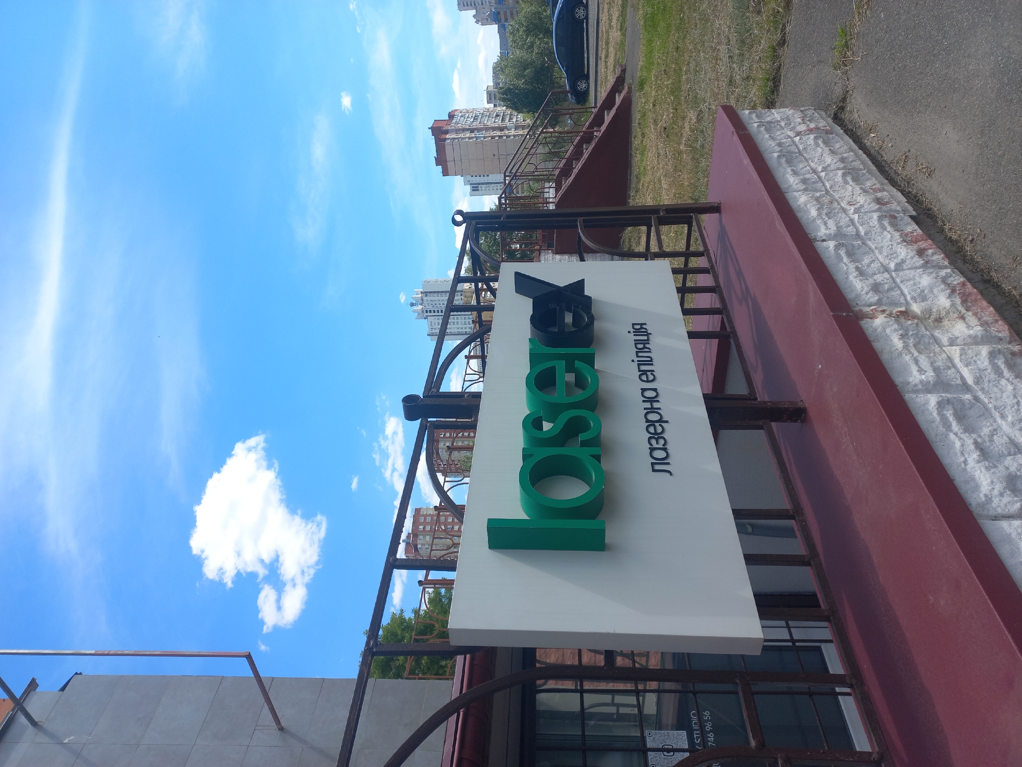 The image displays an outdoor urban scene on a clear, sunny day. The sky is a bright blue with wispy white clouds scattered across it. The photograph is oriented vertically, causing the elements within the frame to appear tilted.
In the immediate foreground, a large white sign is prominently featured. On the sign, vibrant green three-dimensional letters spell out "Laser" (partially obscured, potentially "LaserX"). Below this, in black Cyrillic script, is the phrase "лазерна епіляція," which translates to "laser epilation" or "laser hair removal." Further down, smaller text indicates "STUDIO" and a partially visible phone number, "746 96 56." The sign appears to be part of a business entrance.
The sign is supported by an ornate, dark metal railing or fence structure. Beyond this, a low wall with a reddish-brown base and a top layer of rough, white brick-like stones is visible. To the right of this wall, there's a patch of dry, sparse grass and an asphalt path or road, where a dark car is partially visible. In the background, several multi-story apartment buildings with light-colored facades and some reddish-brown accents rise against the sky, interspersed with green trees.
The scene suggests a commercial area or residential street in a city where Cyrillic script is used, such as Ukraine. The bright sunlight indicates it is daytime, likely midday or early afternoon. No people are visible in the image.