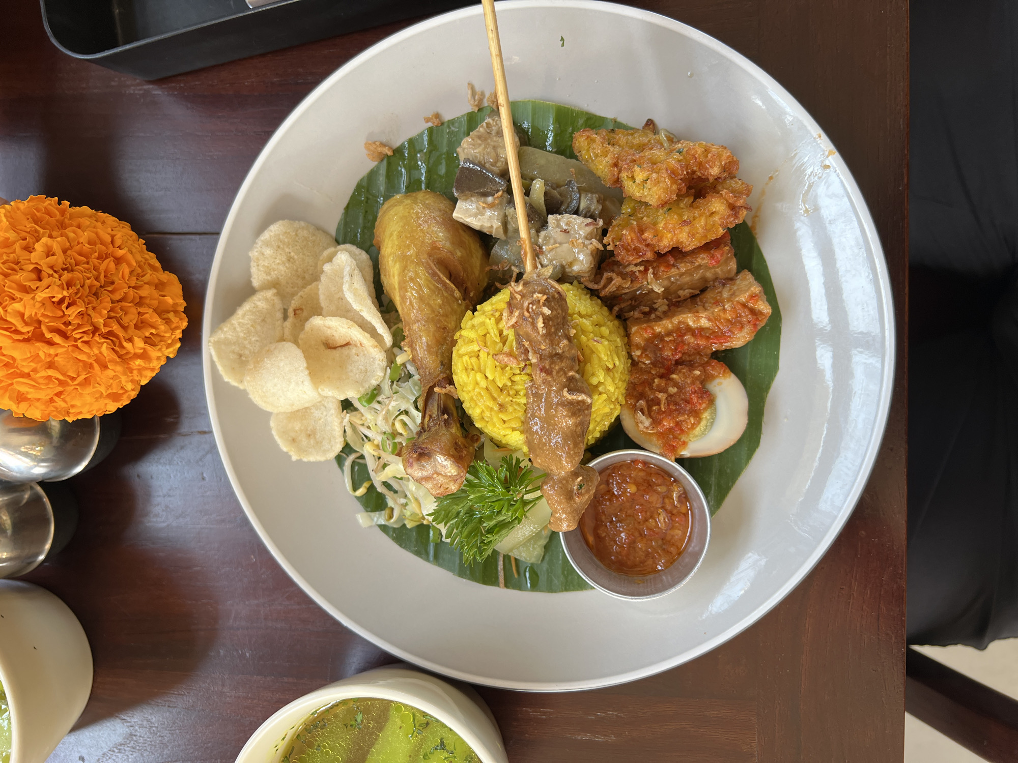 In Ubud District, Indonesia, a close-up, top-down shot captures a plate of Indonesian food, likely from a restaurant, resting on a dark wooden table. The meal features a mound of bright yellow rice, garnished with a skewer of meat. Alongside the rice are pieces of fried chicken, a hard-boiled egg topped with chili, fried tempeh, and what appears to be some sort of vegetable or tofu dish. Crackers and a small bowl of red chili sauce complete the presentation on a banana leaf. To the left, a white bowl holds a green soup, possibly a broth with herbs. An orange marigold, a common sight in Bali, sits in the upper right corner, adding a splash of color to the scene. The overall impression is a vibrant and appetizing meal, ready to be enjoyed.