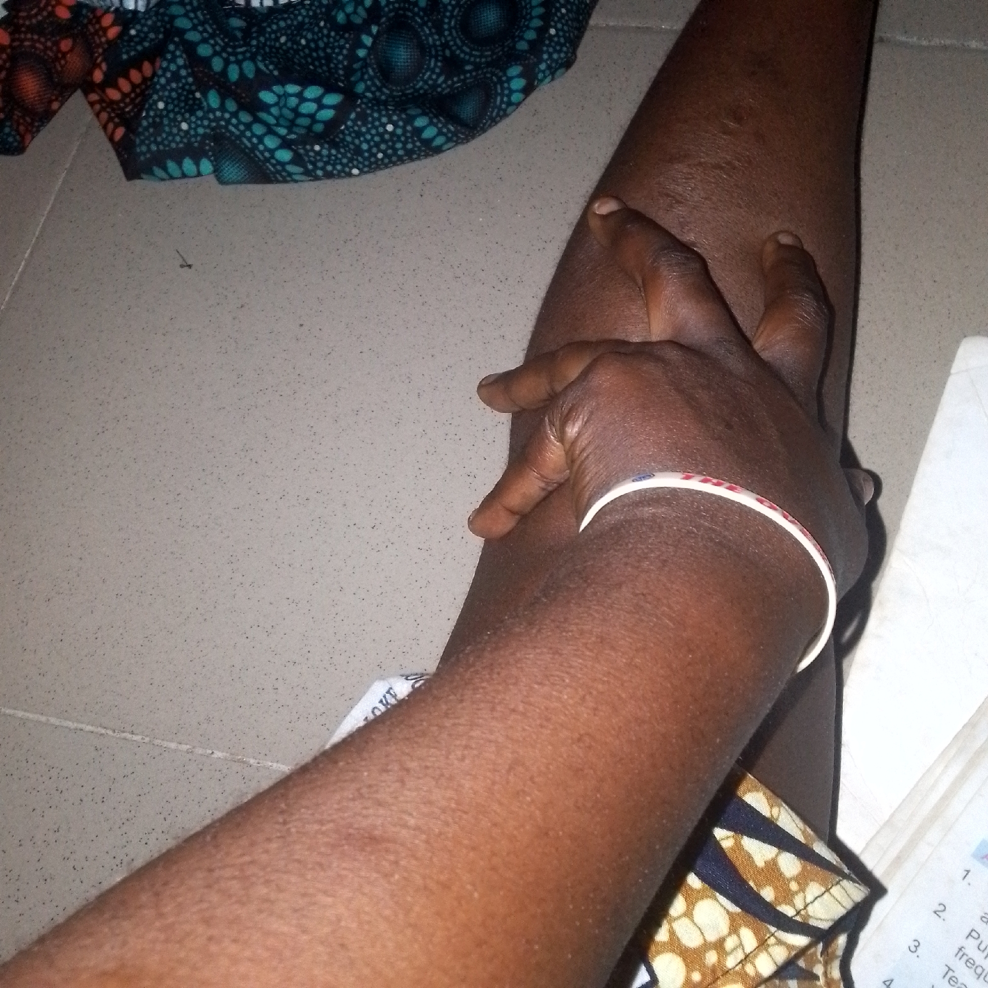 A close-up, top-down shot focuses on a person's dark-skinned arm and hand resting on a speckled, light-colored surface, possibly a floor. The arm is bent, and the hand is positioned to touch the arm, with fingers gently pressing into the skin. A white bracelet with a thin red stripe adorns the wrist. In the background, colorful fabric with geometric patterns is visible, along with a portion of a book or paper with numbered text. The overall scene is indoors, with neutral lighting casting subtle shadows.