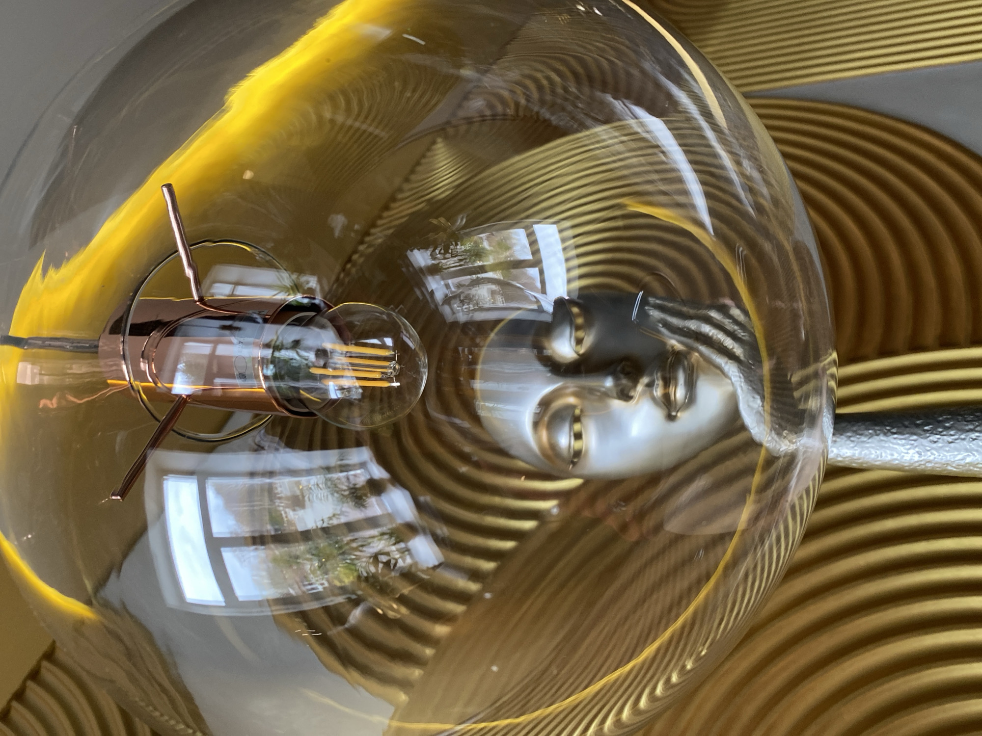 A close-up shot shows a large, clear glass lightbulb with a visible filament inside, set against a backdrop of gold-colored, wave-patterned panels. The lightbulb reflects an exterior scene, including a window and vegetation, and a distorted view of another lightbulb. The location is implied to be Yekaterinburg, Russia.