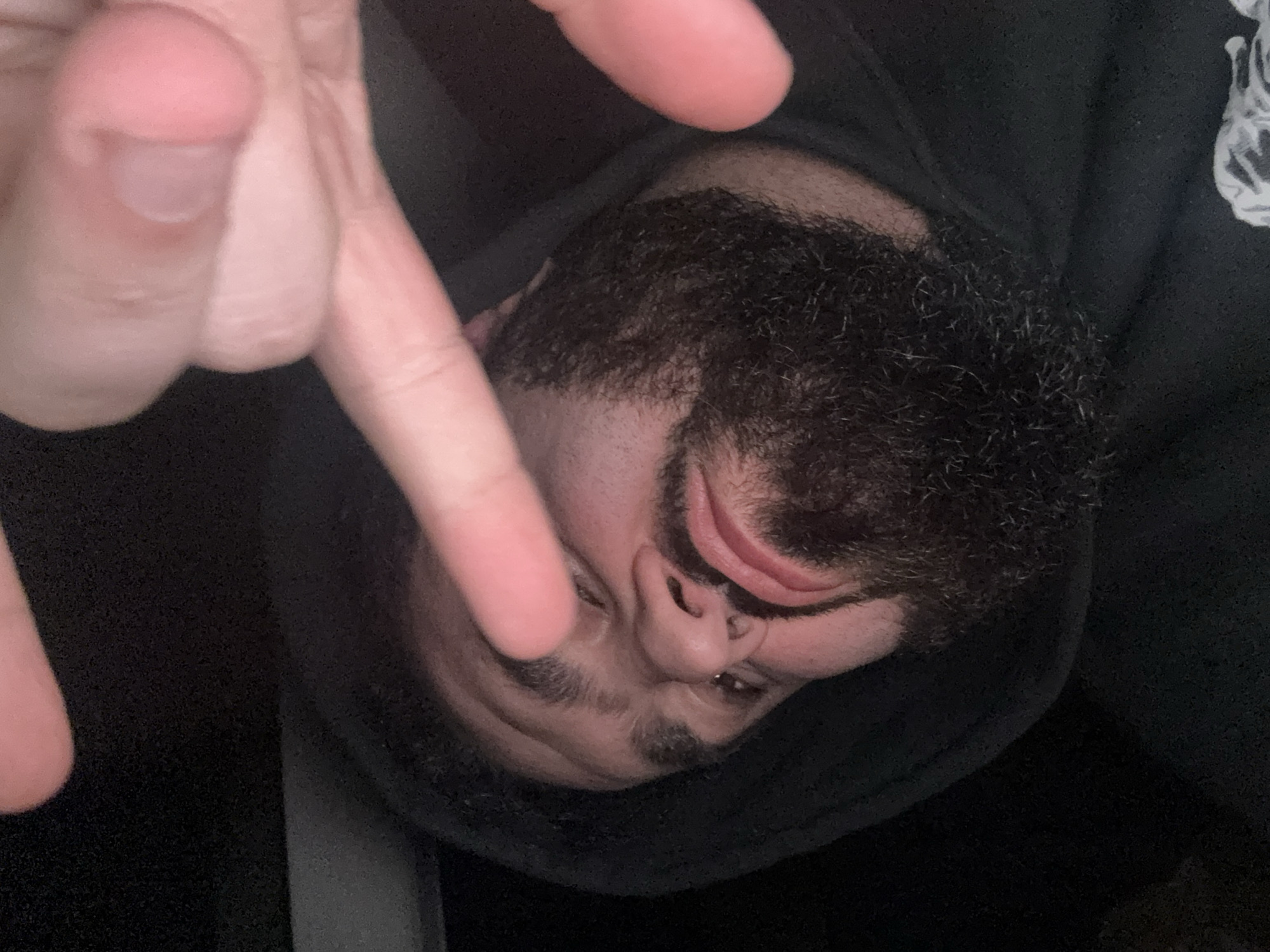 The image depicts a person's face close-up, rotated counter-clockwise. The person has dark hair, a beard, and is wearing a dark hooded garment. A hand with extended fingers is positioned in front of the face. The background is dark and out of focus. No information in the image allows for the determination of its location as Yerevan, Armenia.