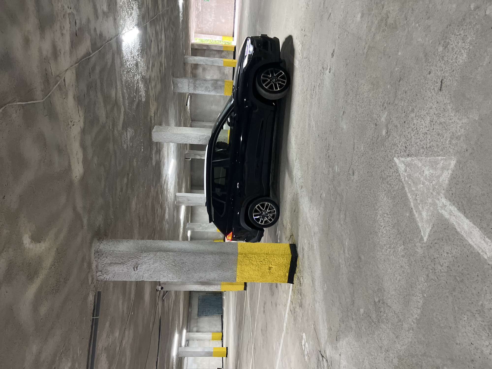 The image shows a black SUV parked in an underground parking garage. The garage has a concrete floor and ceiling, with several concrete pillars supporting the structure. Some of the pillars are painted yellow at the bottom. There are fluorescent lights on the ceiling. White lines on the floor indicate parking spaces, and a white arrow is painted on the floor, pointing forward. The car is positioned in a parking spot to the right of the frame. In the background, there are more pillars, parking spaces, and what appears to be a corrugated metal barrier. The scene is well-lit by artificial lighting. There are no people visible in the image. The location of the image is not determinable from the visual information provided.