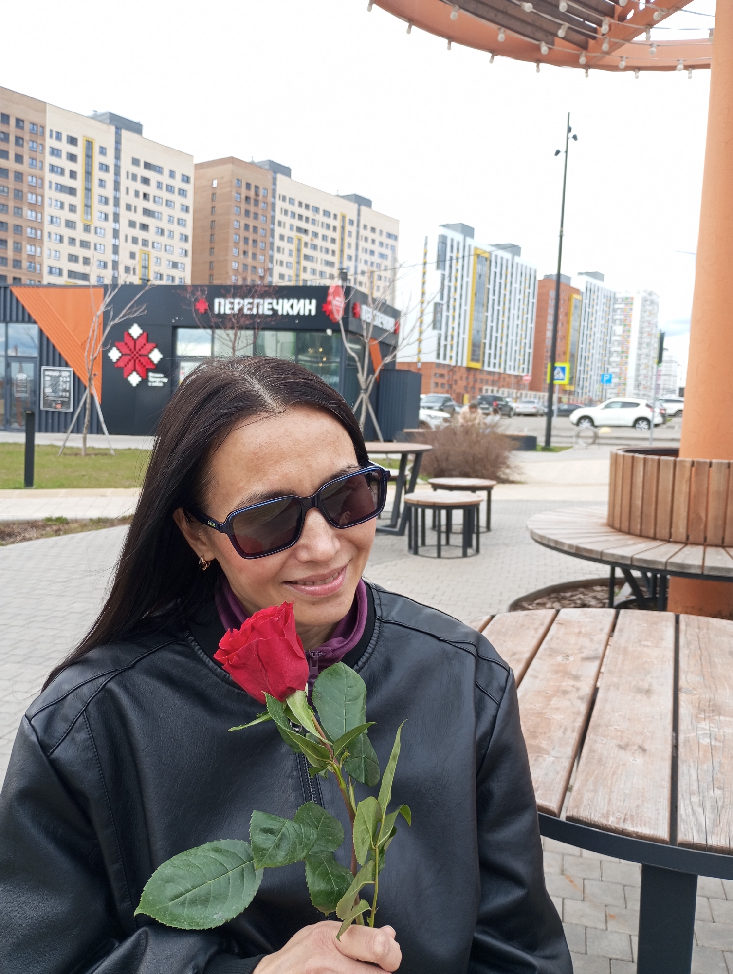 A woman with long, dark hair and dark sunglasses is pictured outdoors, smiling gently while holding a single red rose with green leaves. She is wearing a black leather jacket over a purple top and appears to be seated at a round wooden table in an urban plaza or outdoor seating area with paved ground.
In the background, several modern multi-story buildings of varying colors and styles (beige, white, grey, orange) are visible. Directly behind her, a black commercial building features a striking red and white geometric pattern and the name "ПЕРЕПЕЧКИН" (PEREPECHKIN) in red Cyrillic script above its entrance. Further back, a street with parked cars, a light pole, and more buildings can be seen. The sky is bright and overcast, indicating daytime. The scene conveys a moment of quiet contentment in a contemporary urban setting in Pervomaysky, Russia.