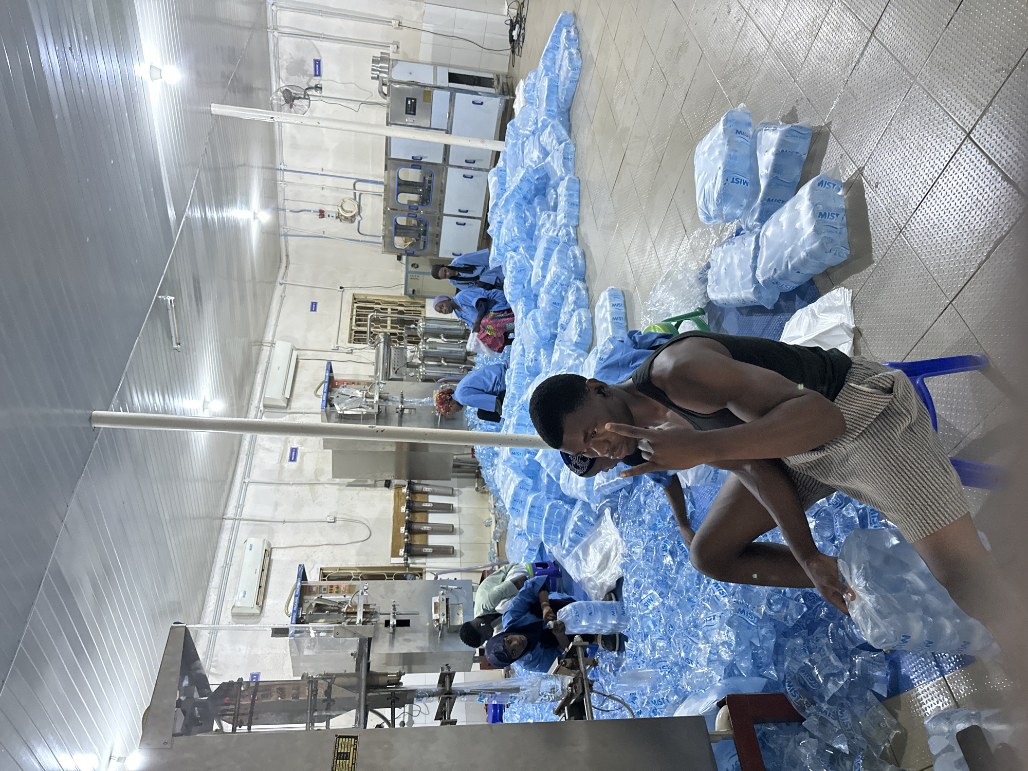 The image depicts the interior of what appears to be a water bottling or packaging facility, potentially located in Jos, Nigeria. Numerous clear plastic bags, labeled with "MIST", presumably containing bottled water, are strewn across the floor and stacked along the production line. The facility has industrial machinery and equipment for water processing and packaging. Several workers are present. One worker, dressed in a black tank top and striped shorts, is seated on a blue plastic chair amidst the bagged water. They are gesturing with their hands, possibly posing for the photograph. Other workers, wearing blue uniforms and head coverings, are positioned near the machinery and bagging areas, seemingly engaged in the packaging process. The environment is utilitarian, featuring metal walls, industrial lighting, and a tiled floor.
