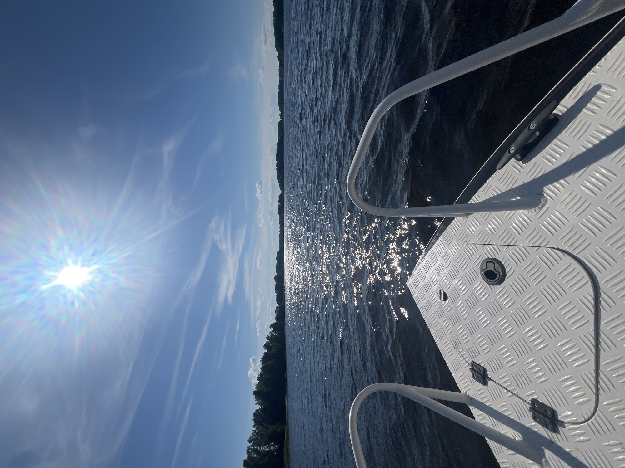 This is a slightly tilted, eye-level, outdoor, medium shot of a sunny day on a body of water, likely a lake, in Сюктерка, Russia. The sun is bright and low in the sky, casting a strong glare on the water's surface and creating a lens flare in the sky. The sky is a clear blue with wispy white clouds. The water is dark and choppy, with numerous bright reflections of the sun dancing on its surface.
In the foreground, the edge of a white boat is visible, featuring a diamond-plate metal deck and a white metal railing. A black cleat is mounted on the deck, and a small, hinged compartment with a circular latch is also visible. The railing curves away from the viewer, suggesting movement or the shape of the boat's bow.
In the distance, a dense line of dark green trees borders the water, indicating a forested shoreline. The overall scene evokes a sense of peacefulness and the enjoyment of a warm, sunny day on the water. The time of day appears to be late morning or early afternoon, given the sun's position and the brightness of the light. There is no visible text in the image.