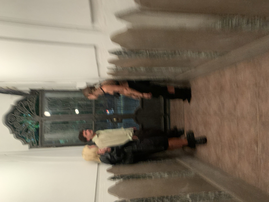 A motion-blurred, eye-level photograph depicts three individuals standing in a narrow, tiled passage within the Recoleta Cemetery in Buenos Aires, Argentina, at night. The passage's walls are lined on both sides by repetitive, grey, pointed structures resembling small tombstones or decorative grave markers. At the end of the passage, a tall, dark metal door or large window with ornate scrolled ironwork at the top reveals faint exterior lights.
From left to right: a blonde-haired individual, dressed in a dark jacket and dark lower garments, is partially obscured by the left wall. A male individual with dark hair and a mustache, wearing a light-colored long-sleeved shirt and dark pants, stands facing right. To his right, a female individual with dark hair, wearing a dark bra-like top, dark pants or skirt, and dark boots, stands facing left. The central male and the female on the right appear to be engaged in a casual conversation. The floor is covered in reddish-brown tiles, and the upper portions of the walls are white.