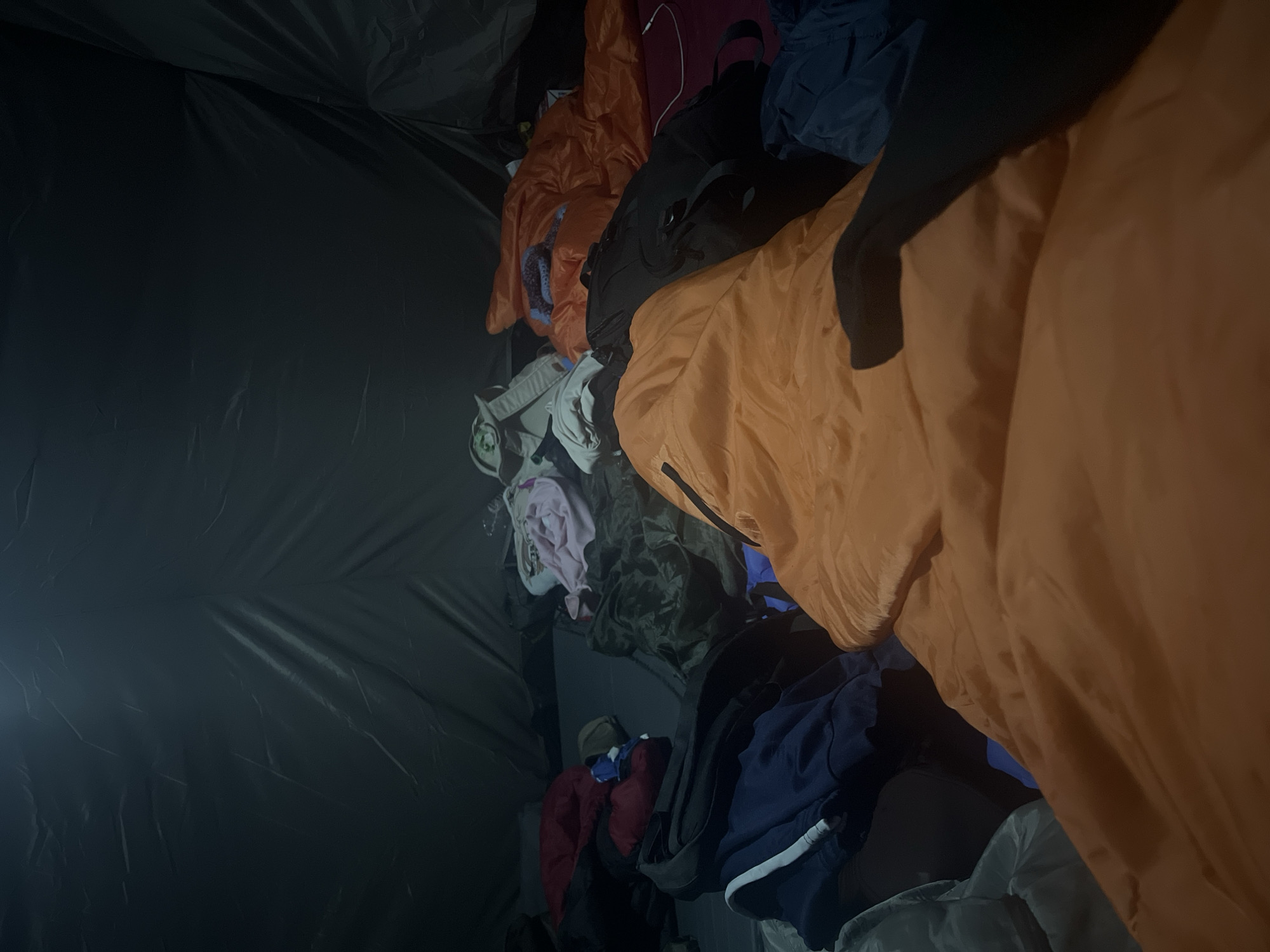 The image appears to be taken inside a tent or enclosed space with a dark, possibly black plastic or tarp-like material forming the walls. A pile of clothes is visible, consisting of various colors and textures. There are items in shades of orange, blue, red, black, pink, and gray. Items appear to include jackets, hoodies, and possibly other clothing items. There's also a glimpse of what might be a white cable. The lighting is dim, with a light source visible on the left side of the frame. No people are directly visible. The location of Lesnoye, Russia, is not directly verifiable from this image.