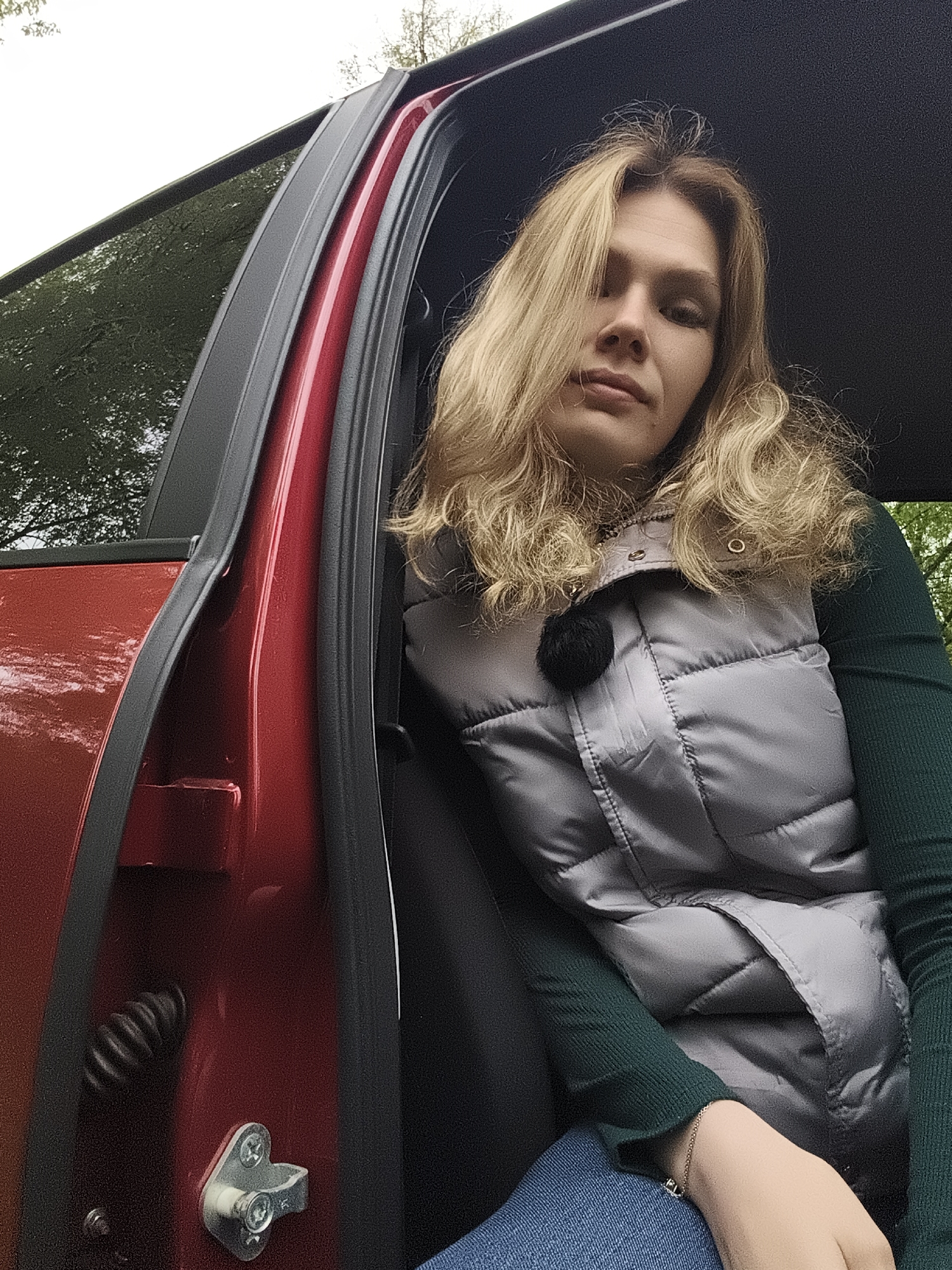 A woman with blonde, shoulder-length curly hair sits inside a red car. She wears a gray puffy vest over a dark green long-sleeved shirt and blue jeans. The car door is open, revealing a black interior. A metal latch and a coiled spring are visible on the red car door frame. The background shows trees and a hint of daylight, suggesting an outdoor setting. There is no information present in the image to determine the location in the city of Izhevsk, Russia.