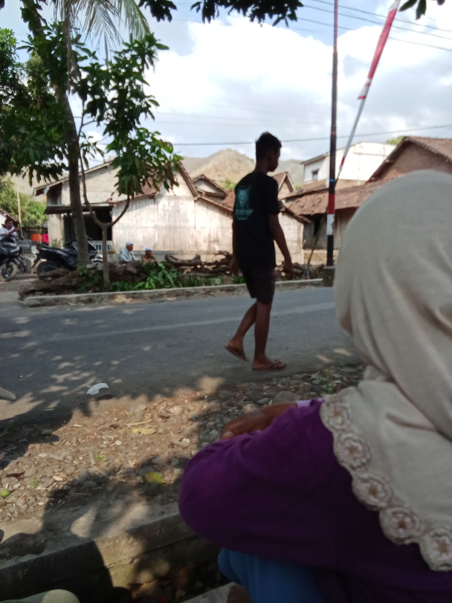 In the city of Trebungan, Indonesia, a casual scene unfolds on a roadside. A man, dressed in a black t-shirt and shorts, walks along the asphalt. In the foreground, the back of a person wearing a light-colored hijab and a purple garment is visible, suggesting they are observing the street. The environment is typical of a village in Indonesia, with simple houses and trees lining the road. The sky is a bright blue with scattered clouds, indicating a sunny day. The overall impression is one of a quiet, everyday moment captured in a small Indonesian town.