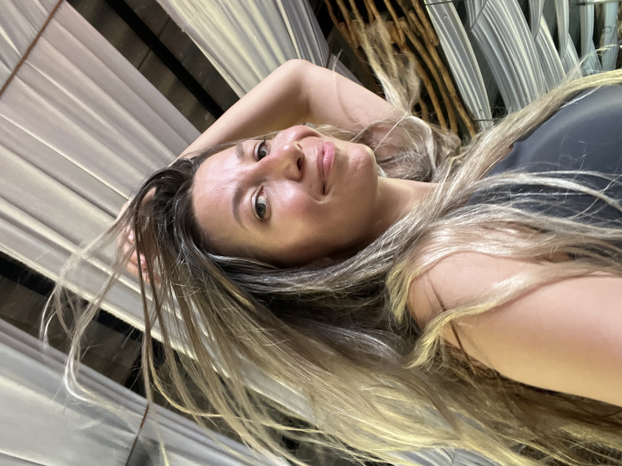 A woman with long, blonde hair is looking at the camera and smiling. Her hair is windblown and falling around her face. She is wearing a dark-colored top. Her right arm is raised and resting on her head. The background consists of fabric draped from a ceiling, creating a tent-like effect, with wooden beams visible behind the fabric. The lighting suggests an indoor setting, possibly with natural light entering from an unseen source. The overall impression is casual and candid.