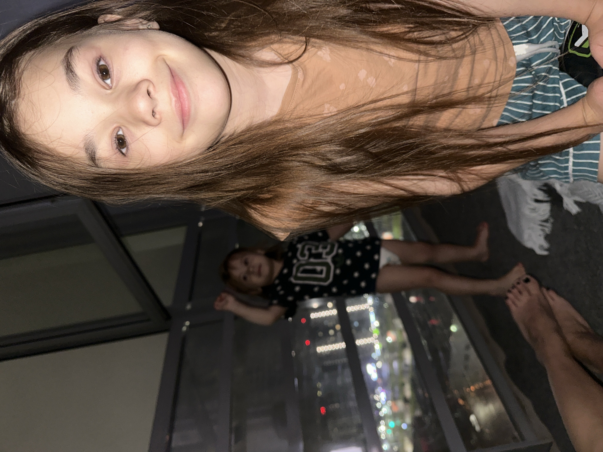 The image captures a playful moment with two individuals, likely siblings, positioned on an upper floor overlooking a city at night. The primary subject, a young girl with long, dark brown hair and a light complexion, looks directly at the camera with a slight smile. She wears a light brown or tan top with subtle light-colored patterns and a striped blue and white bottom. An earring is visible in her left ear.
In the background, a smaller child, possibly a toddler, stands further back, also looking towards the camera. This child wears a dark t-shirt with a white number "50" on it and a light-colored diaper or shorts. Both children are barefoot, and the feet of an adult are visible in the lower right corner, suggesting another person is present.
The scene appears to be indoors, against a large window or glass wall that offers a panoramic view of a city illuminated by countless blurred lights, indicating nighttime. The artificial lights suggest a lively urban environment. The reflections on the glass hint at the indoor setting. The overall mood is casual and relaxed, capturing an intimate family moment. There is no visible text in the image other than the number on the child's shirt. The location is specified as Izhevsk, Russia.
