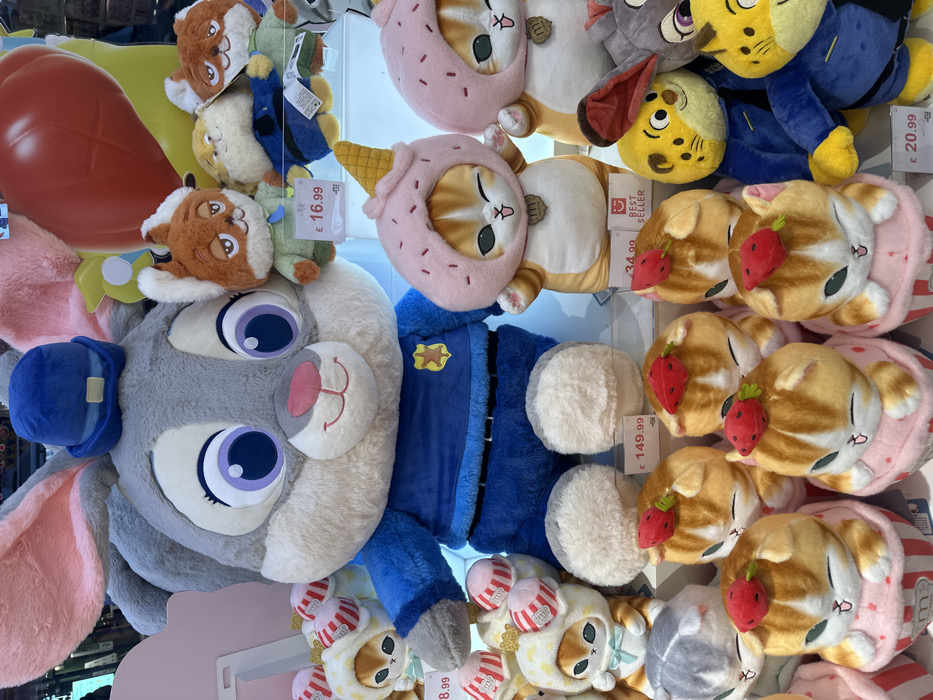 A collection of plush toys in a store window. Judy Hopps from Zootopia is prominently featured. Several cat plushies with strawberry and ice cream cone designs are displayed. Other characters include a fox and a yellow tiger. Price tags are visible, indicating the cost of some items. The toys are densely packed and brightly colored, creating an appealing display.