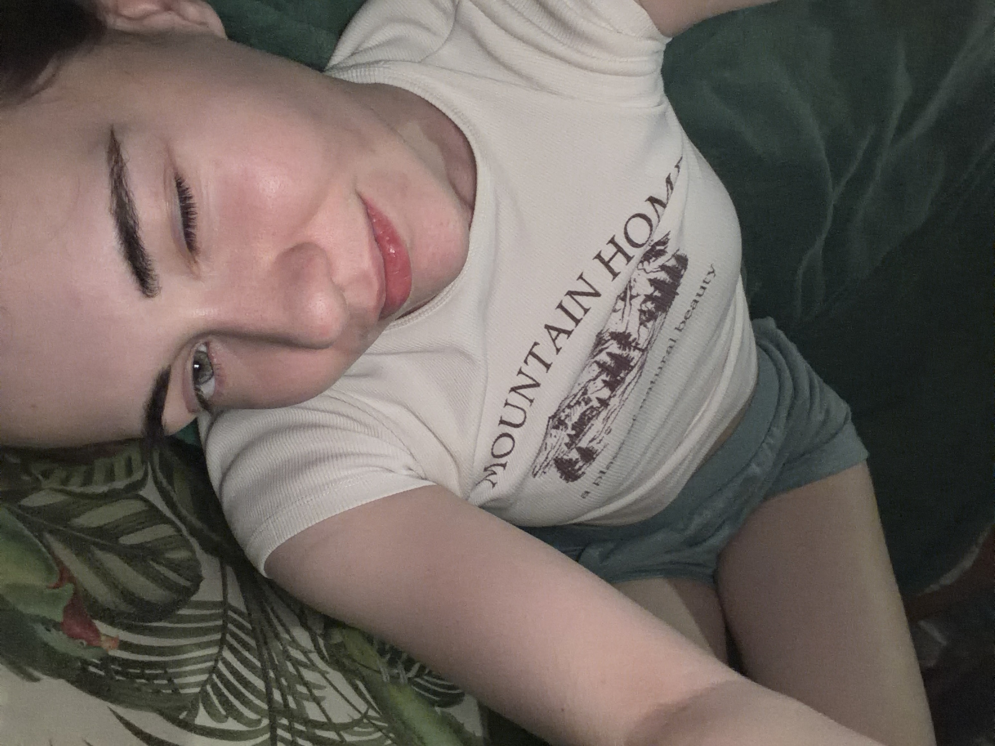The image shows a person lying down, with their head tilted. They are wearing a white t-shirt with "MOUNTAIN HOME" and a graphic of mountains printed on it, and blue shorts. The person's left eye is open and looking at the camera, while their right eye is partially closed. Their lips are slightly parted and appear to be glossy. The background is a dark green textured surface, possibly a blanket or couch, and a pillow with a leaf pattern. The image is taken from a high angle, looking down at the person. There is no information in the image to suggest a location in Zavodoukovsk, Russia.