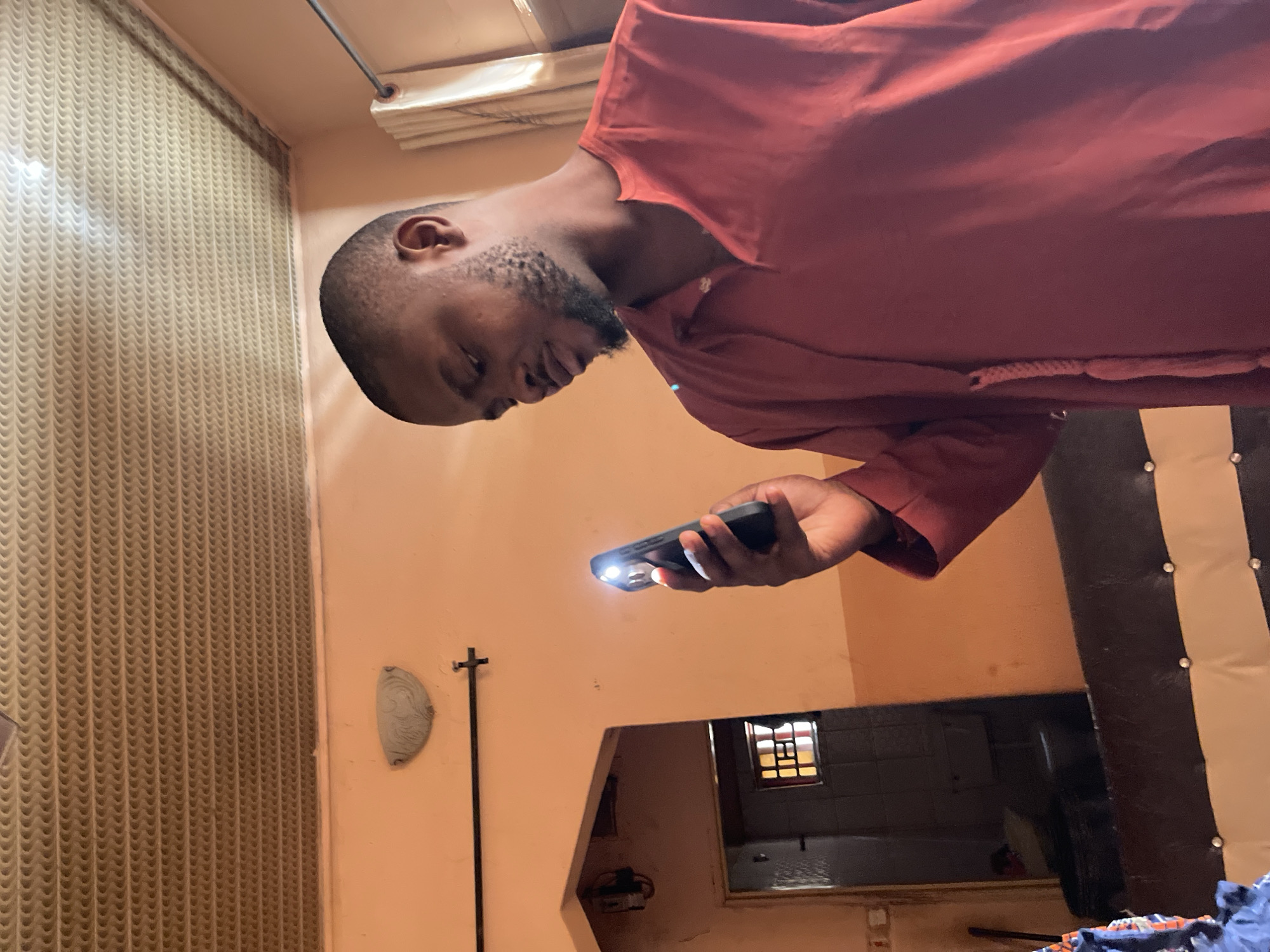 A man with a beard, wearing a red shirt, is holding a smartphone with its flashlight on. The scene is indoors; wallpaper is visible on one wall. A mirror reflects a bathroom with a window and tiling. The location is likely Muchiya, Nigeria, based on the user's specification.