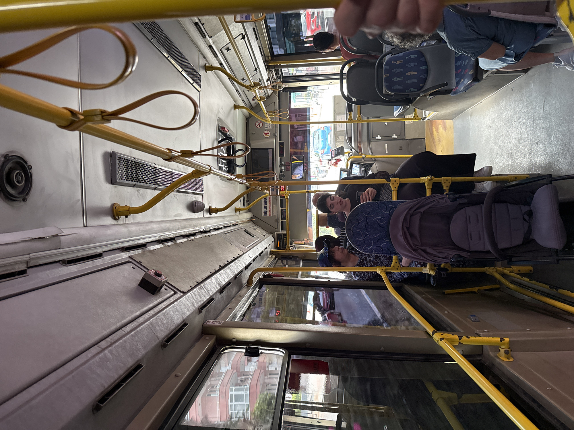The image depicts the interior of a public transit bus in Alanya, Turkey, viewed from a slightly elevated position near the front, looking towards the rear. The bus interior features a light grey ceiling with integrated air vents, a central lighting strip, and multiple yellow horizontal grab bars with brown leather-like hanging straps. A circular speaker is visible on the ceiling. The walls are lined with light grey-brown panels and large rectangular windows. The floor is covered with worn grey non-slip material.
On the right side, near the front, a sign in Turkish reads "İNİŞLER SADECE ARKA KAPILARDAN!" (Exits only through rear doors!). A red "DUR" (Stop) button is mounted on a black box attached to the ceiling panel on the right. Small display screens are visible above the front window area. Yellow railing systems extend along the windows and divide the seating areas from the central aisle.
Several passengers are visible. In the front-left, a woman with dark hair, wearing a black top, is seated and looking down at a smartphone in her hands. A patterned bag rests on her lap. Adjacent to her is a dark purple/grey baby stroller. Further back on the left, a woman wearing a dark patterned top and a blue baseball cap is seated, looking towards the left. On the right side, further towards the rear, a person with dark hair in a red shirt is seated. In the far right foreground, a passenger in a light blue denim-like top is partially visible. The seats are grey with a distinctive blue and light-colored pattern depicting small human figures.
Through the bus windows, an urban environment characteristic of Alanya, Turkey, is visible. Multi-story residential buildings with pinkish-beige and white facades and balconies are prominent. A blue sedan is visible on the road through the front windows, and a red car is seen through the right-side window. The street appears to be asphalt, and the overall lighting suggests an overcast day or interior reflections. The Turkish signage confirms the location within Turkey, consistent with the user's provided context of Alanya.