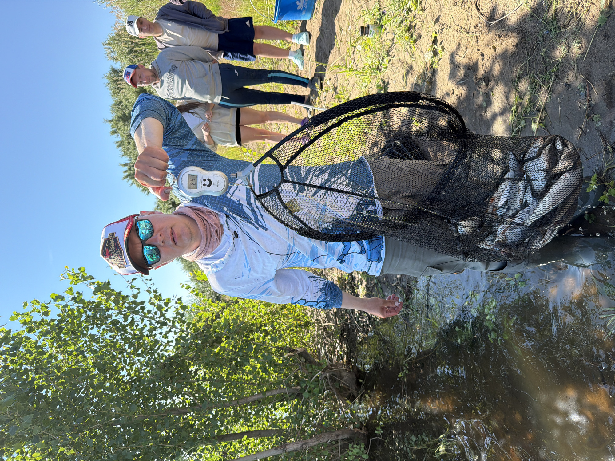 In Tarasovo, Russia, a man in a blue long-sleeved shirt, khaki shorts, and a red and white baseball cap stands by a body of water, holding a fishing net filled with fish. He is also holding a digital scale that reads "6.4". Behind him, three other individuals are visible, along with trees and a bright blue sky. The ground is dirt and grass.