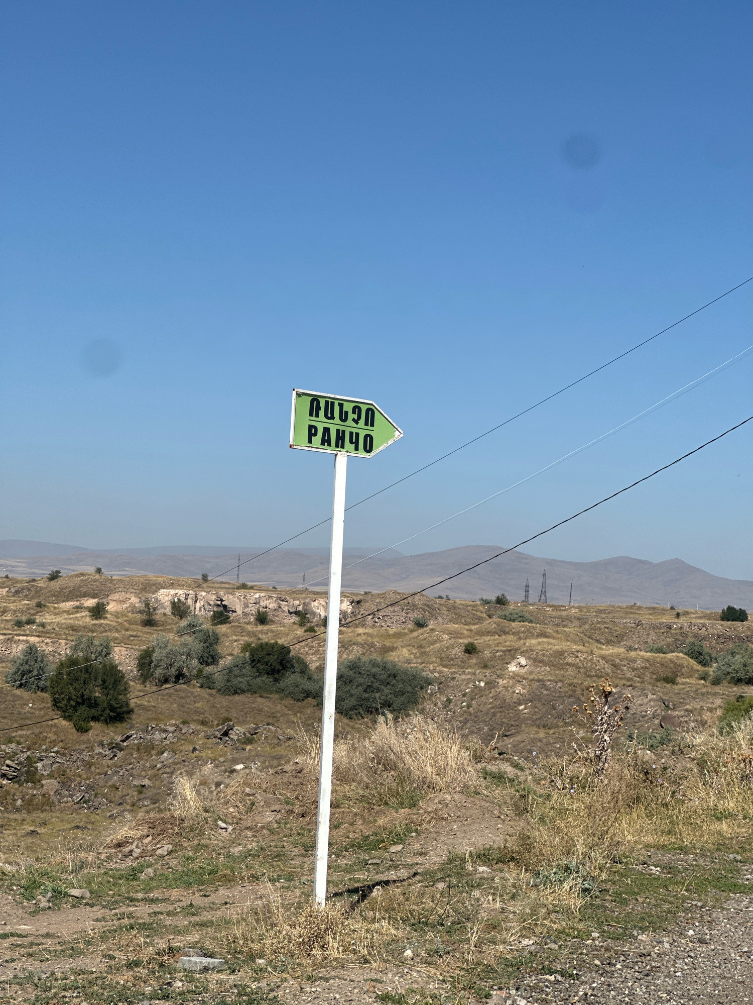 A green, arrow-shaped directional sign points to the right, with Armenian text reading "ՎԱՆՔՈ" and "РАНЧО" printed in black. The sign is mounted on a white pole. The background features a dry, hilly landscape with sparse vegetation, rocky outcrops, and distant mountains under a clear blue sky. Power lines are visible stretching across the scene. The environment suggests a rural or semi-rural area, possibly on the outskirts of Gyumri, Armenia, given the Armenian script on the sign.