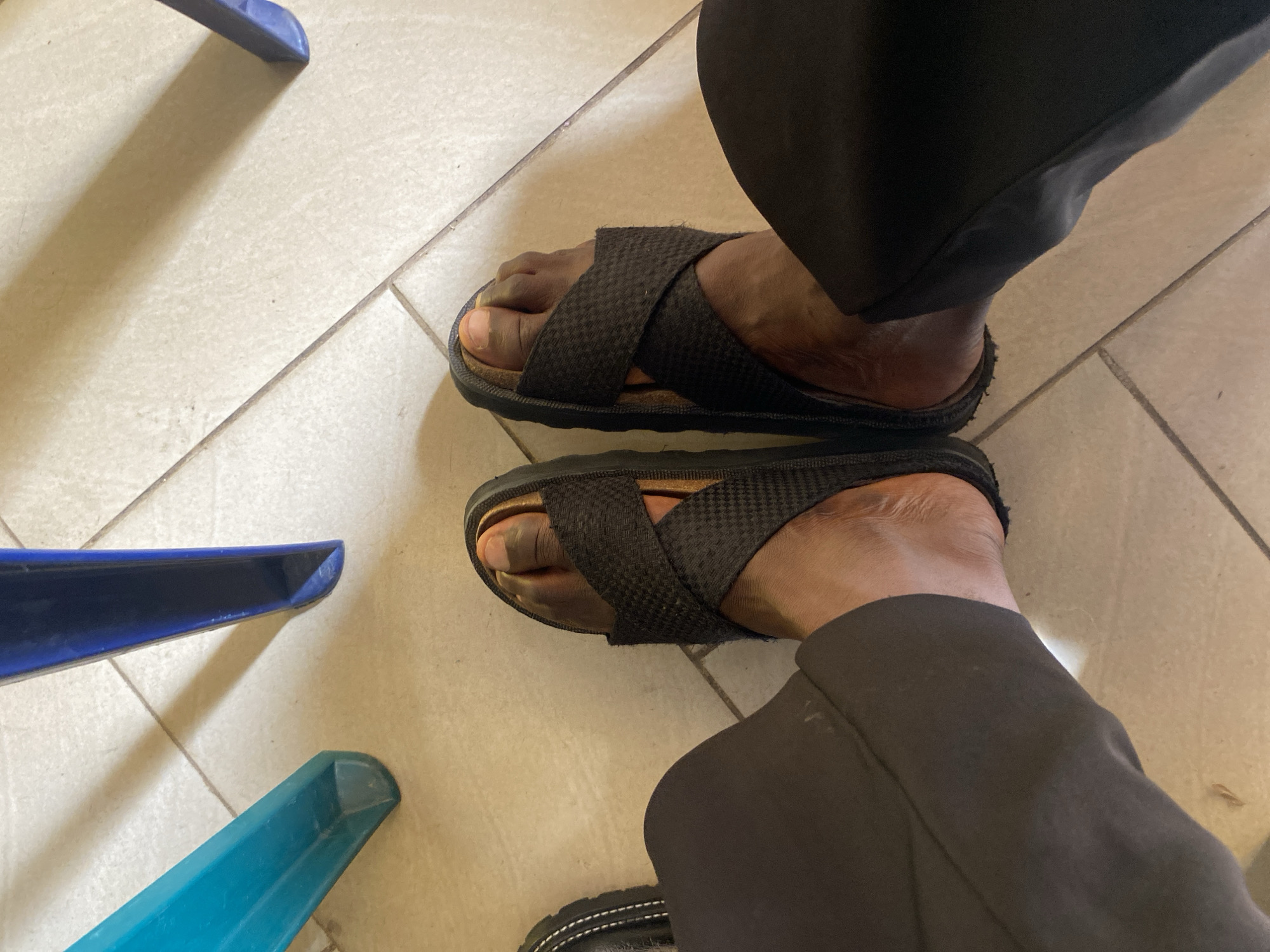 A high-angle shot depicts a person wearing black slides with crisscrossed straps while seated on a chair with blue and teal legs. The person is also wearing black pants. The floor is covered with beige tiles. The location is in Gura Top, Nigeria.