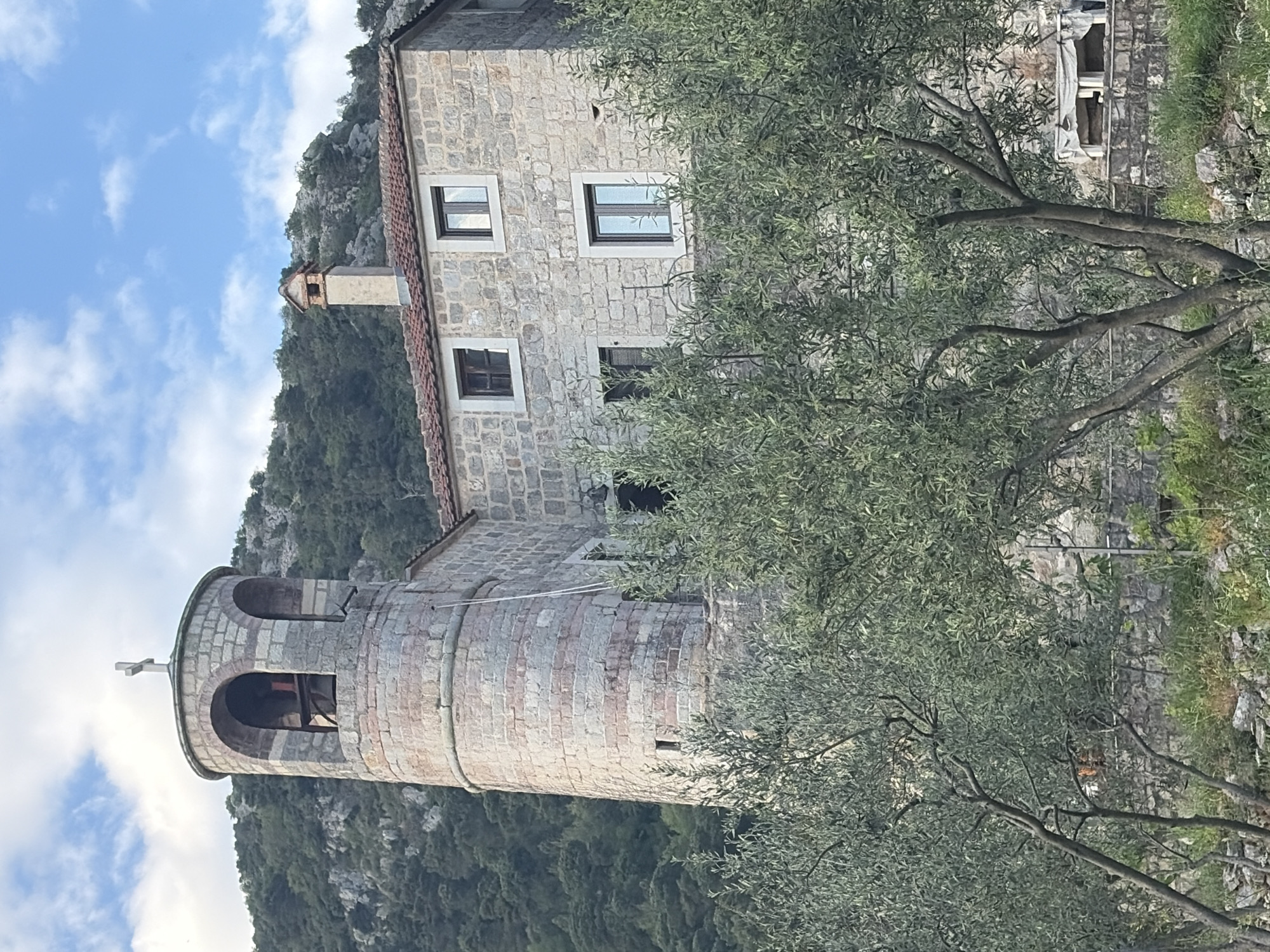 A low-angle view presents a stone building complex in the vicinity of Budva, Montenegro, set against a backdrop of mountainous terrain and a partially cloudy sky. Dominating the left is a cylindrical tower with arched openings near the top, topped with a cross. Adjacent to the tower is a three-story building made of the same stone material, with windows visible on each level and a chimney protruding from the roof. A tree with thin branches and small leaves fills the right side of the frame, partially obscuring the building. The surrounding landscape consists of dense green vegetation covering the mountains.