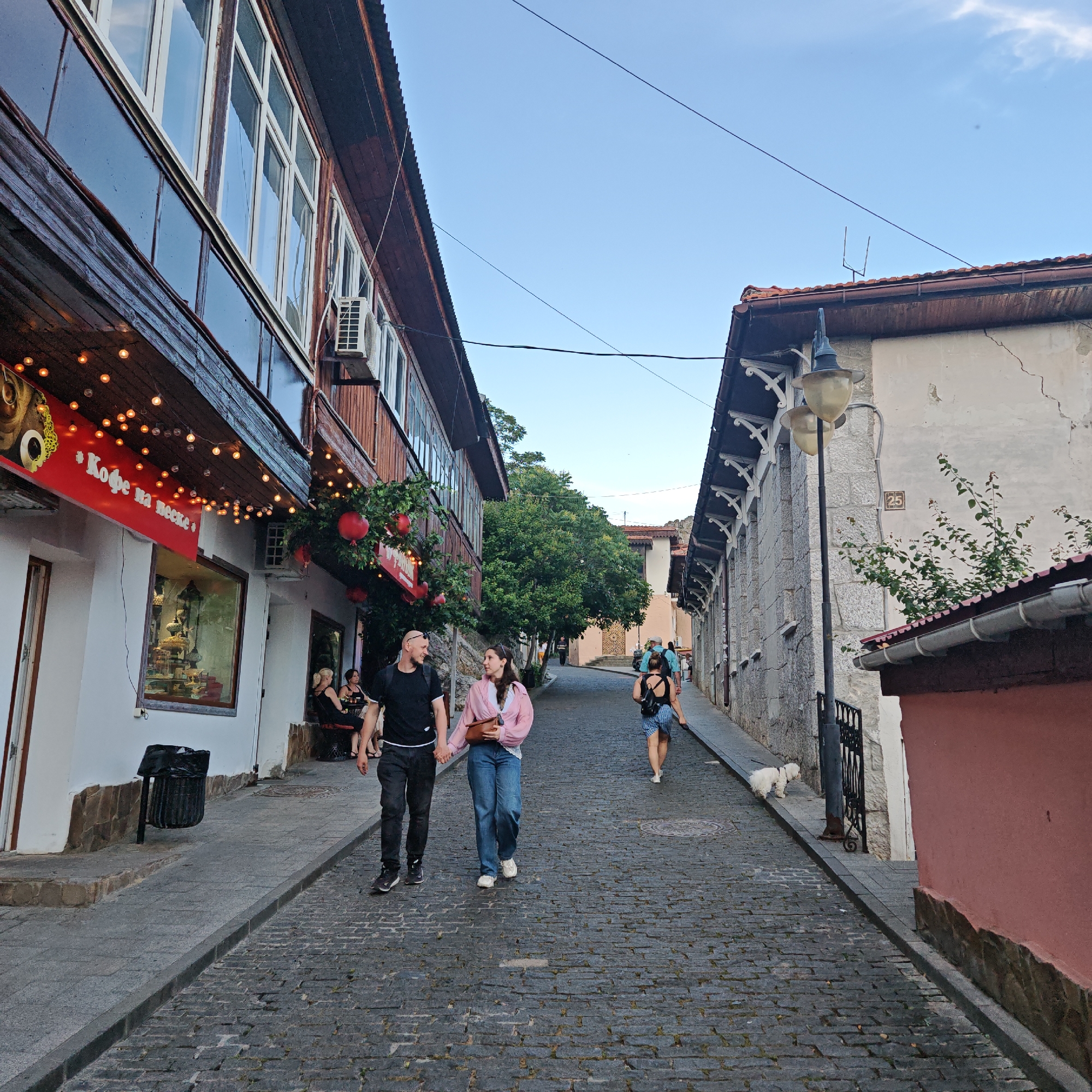 A couple walks hand-in-hand down a cobblestone street lined with buildings in Gurzuf, Ukraine. The building on the left has a wooden facade with large windows and is decorated with string lights and red hanging decorations. A sign reads "Кофе на песке," which translates to "Coffee on the sand." Several people are seated at tables outside this establishment. The building on the right is constructed of stone with white decorative trim under the eaves and a street lamp. Further down the street, more people are walking, and a small white dog is visible on the sidewalk. The sky is clear and blue.