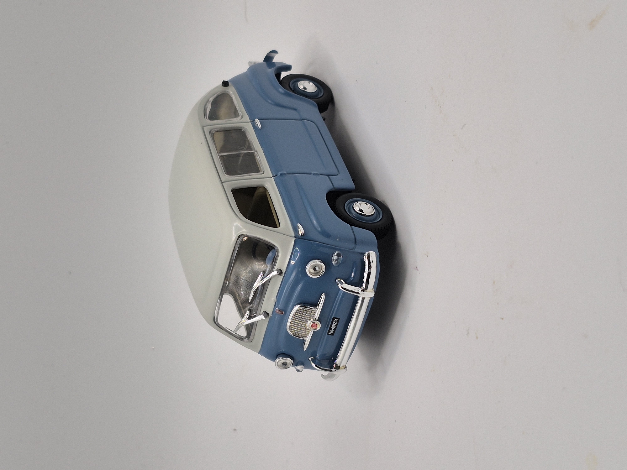 A model of a blue and white two-tone retro van is positioned on a plain white surface, casting a faint shadow. The van is facing slightly to the left of the viewer, with its front grille, headlights, and chrome bumper clearly visible. A license plate on the front reads "NA-A01234". The van has visible side windows and two windshield wipers. The background is a seamless, light gray color, suggesting a studio or controlled environment. There are no people, animals, or other objects present in the image, and the scene does not depict any specific location within the city of Schwäbisch Gmünd, Germany.