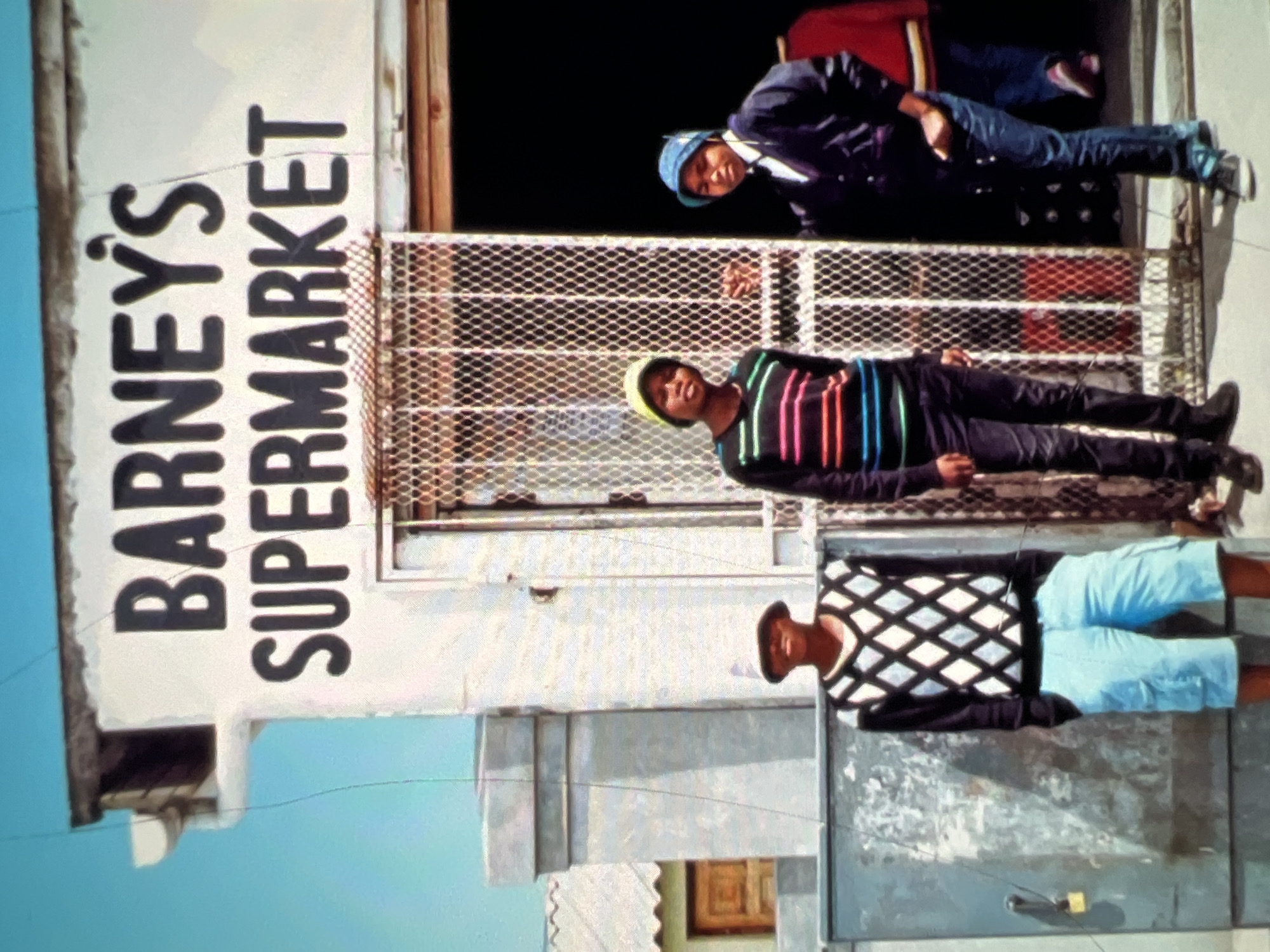 The image depicts a street scene in Lagos, Nigeria, with a storefront named "BARNEY'S SUPERMARKET" prominently displayed. Three young individuals are visible in front of the supermarket. On the left, a person wearing a dark sweater with a white diamond pattern and light blue cargo shorts stands with their hands in their pockets. In the center, another person is leaning against a metal grate, wearing a striped long-sleeved shirt with black, pink, green, and blue stripes, dark pants, and a yellow cap. To the right, a third person, wearing a dark jacket over a white shirt and a blue cap, is standing with one hand in their pocket, facing the camera. Behind them, a portion of another person wearing a red jacket is visible inside the supermarket entrance. The storefront itself appears weathered, with a metal gate covering the entrance. The environment suggests an outdoor setting with a clear blue sky.