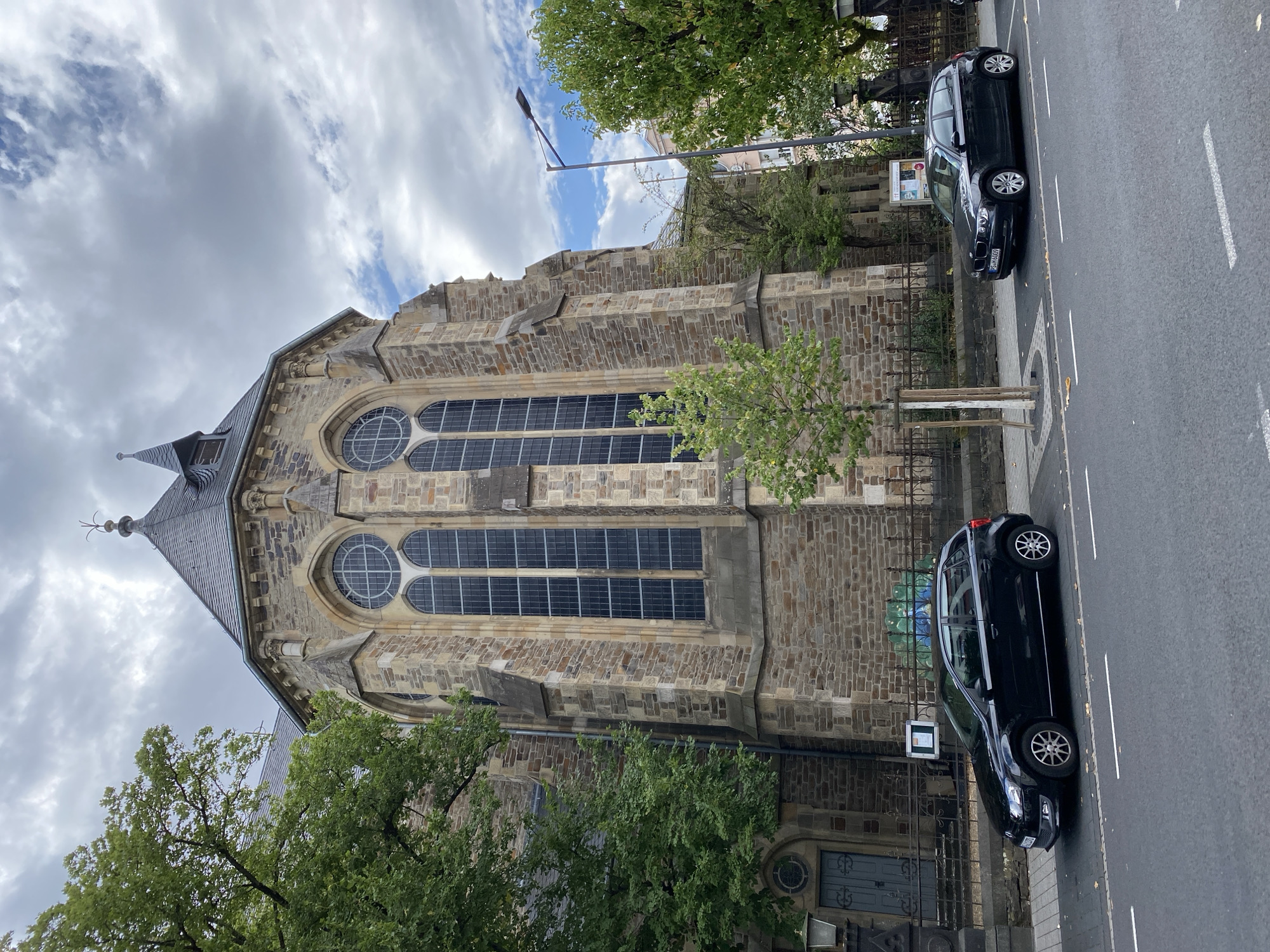 The image depicts the exterior of the Church of St. Matthew in Neuwied, Germany. The church is constructed from stone, featuring a tall, pointed roof and large arched windows with a grid-like pattern. In the foreground, two black cars are parked on a street adjacent to the church. The street is paved and has white lane markings. A sidewalk runs between the street and the church's fence. Trees with green foliage are present on both sides of the church and along the sidewalk. The sky is overcast with clouds, but patches of blue are visible. The scene appears to be a daytime view of the church and its surroundings in a European city.