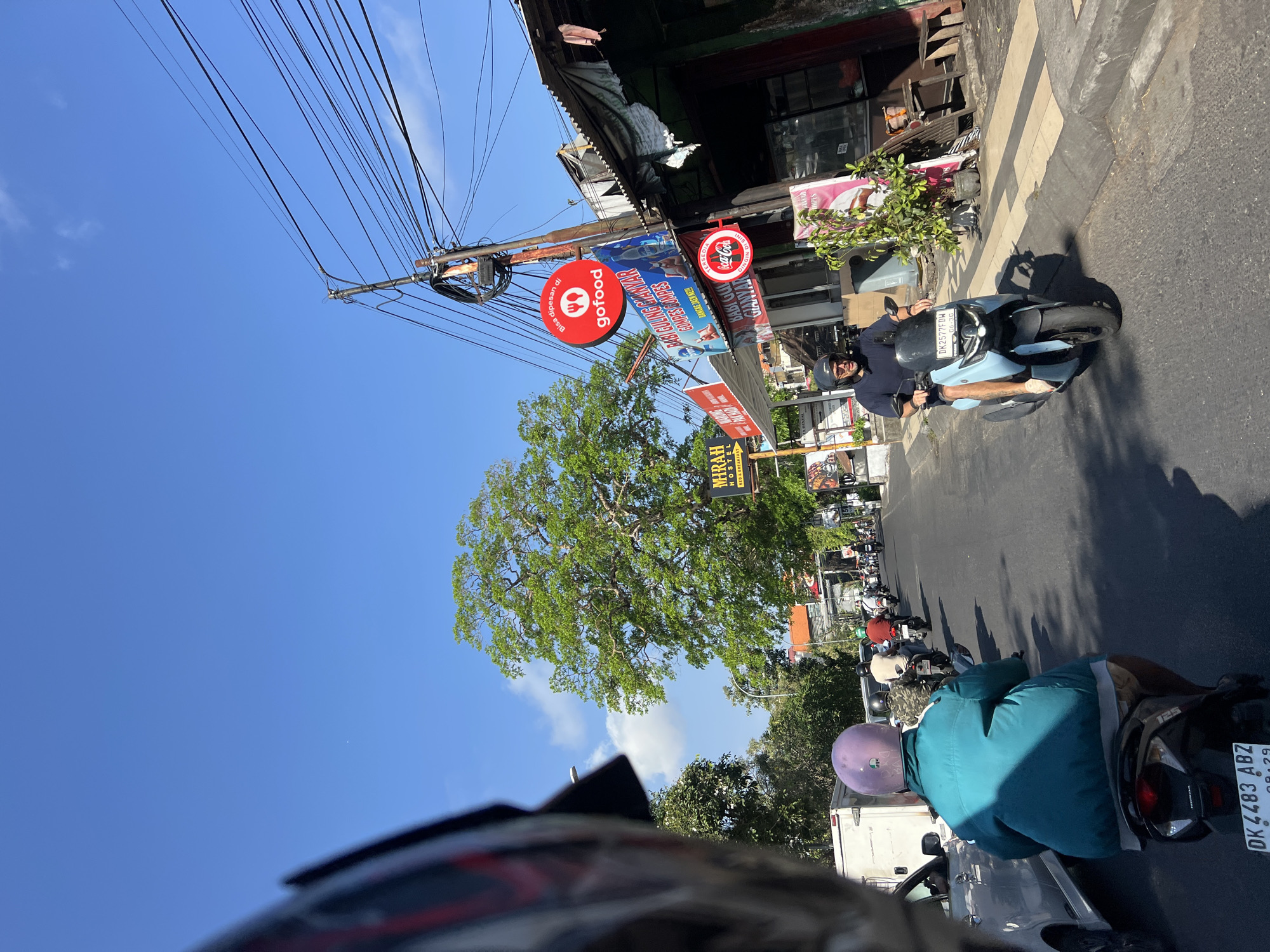 The image shows a street scene in Kuta, Indonesia, on a sunny day. The perspective is from someone riding on the back of a scooter, looking forward. In the foreground, the back of a turquoise jacket and a purple helmet are visible. The scooter has a license plate starting with "DK 4483 ABZ." Ahead, there are other scooters and cars on the road. A man in a dark blue shirt and helmet is riding a light blue scooter in the right lane. To the right of the road, there are buildings with corrugated metal roofs, signs for "Gofood" and "Coca-Cola," and a large green tree. The sky is clear blue with a few wispy clouds. The road appears to be a busy commercial area.