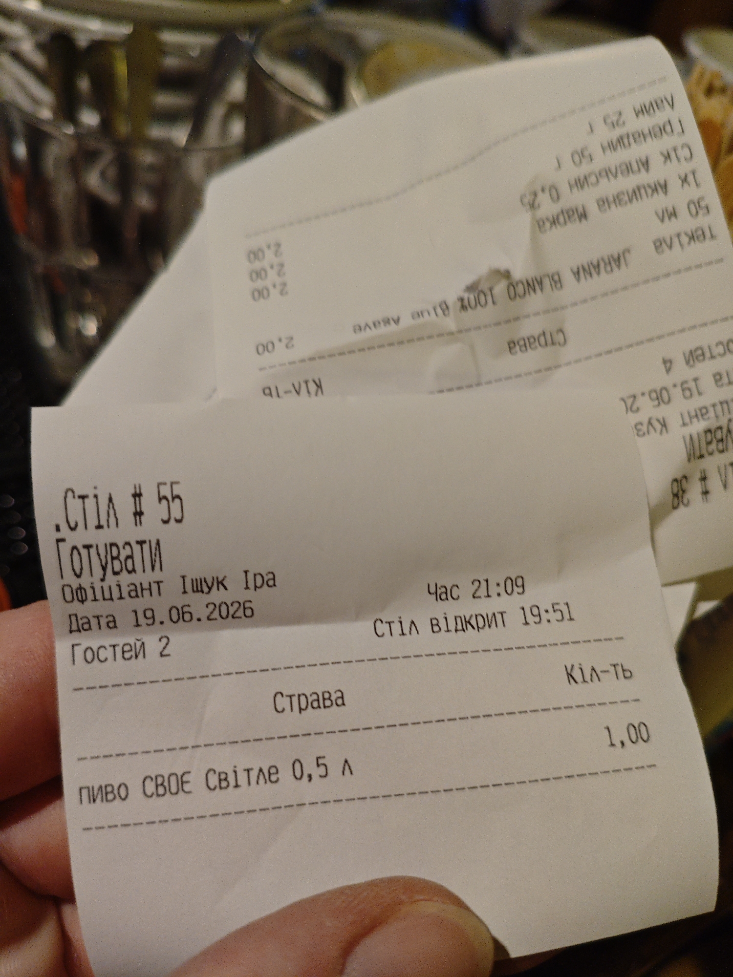 The image shows a close-up of a receipt with Ukrainian text. The receipt is held by a person's hand, with a blurred background that appears to be a restaurant or bar setting. The text on the receipt is in Ukrainian and includes items like "Стіл # 55" (Table # 55), "Готувати" (To cook), "Офіціант Іщук Іра" (Waiter Ishchuk Ira), "Дата 19.06.2026" (Date 19.06.2026), "Гостей 2" (Guests 2), and "пиво СВОЄ Світле 0,5 л" (beer VOE Light 0.5 L). It also shows times like "Час 21:09" (Time 21:09) and "Стіл відкрит 19:51" (Table opened 19:51). There is also another partial receipt visible behind the main one with more items and prices, written in Cyrillic script and rotated. The image does not contain any visual cues or landmarks that would allow for pinpointing a specific location within Kyiv, Ukraine.