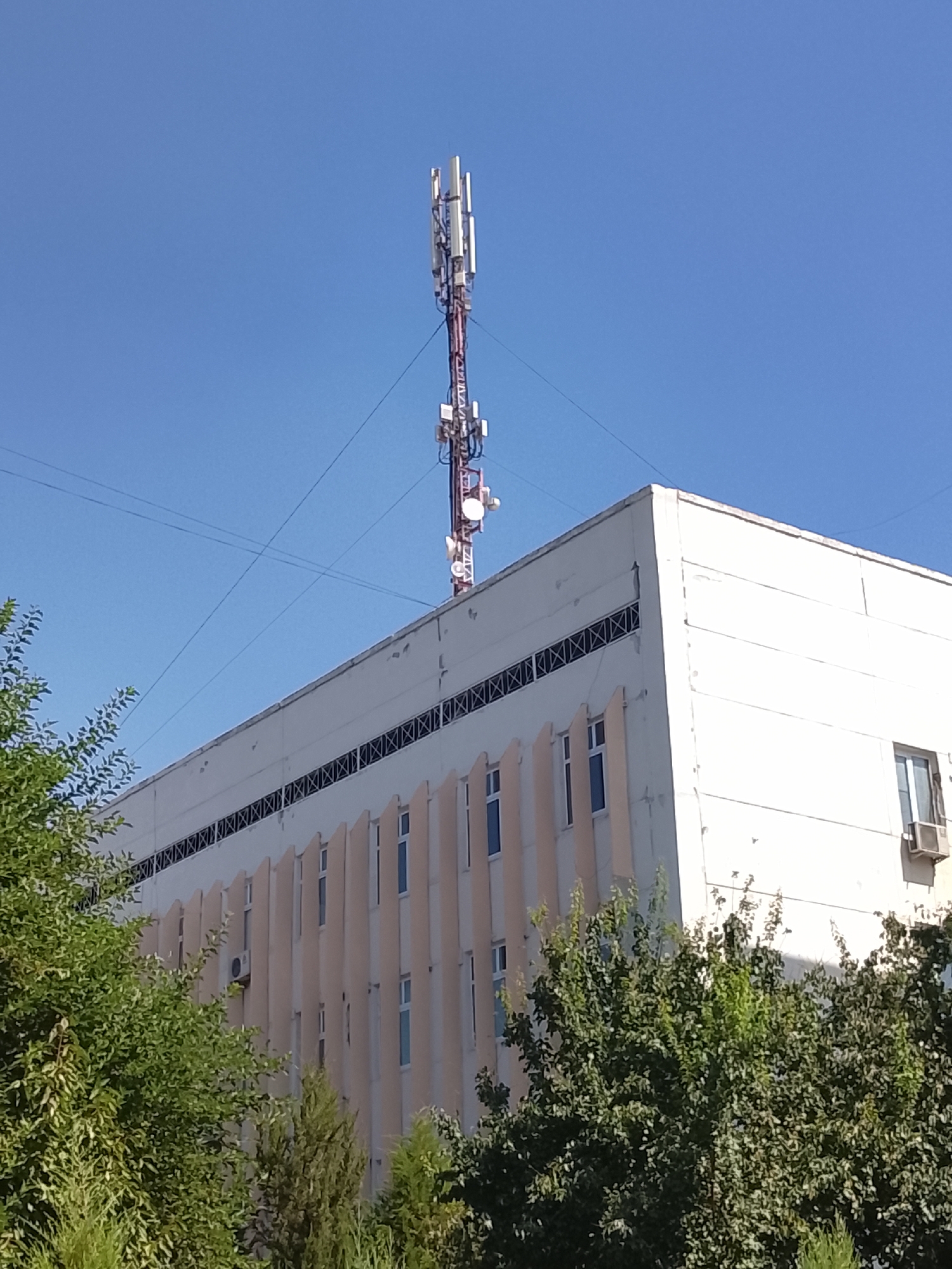 A cell tower stands tall against a clear blue sky atop a cream-colored, multi-story building. The building features a row of decorative panels running along its facade, interspersed with regularly spaced windows. Some of these windows have air conditioning units mounted beneath them. Trees with lush green foliage partially obscure the lower portion of the building. The image appears to have been taken in Fergana City, Uzbekistan.