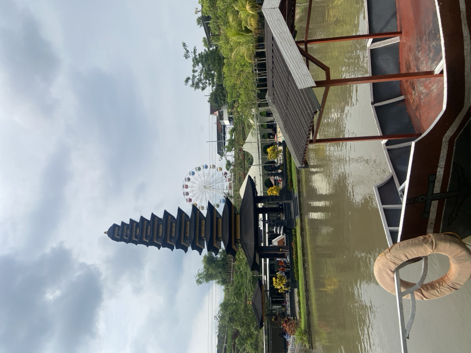 A multi-tiered temple stands prominently against a cloudy sky with a Ferris wheel visible in the distance. Lush greenery flanks the temple and lines the water's edge. A portion of a boat with a brown and white hull is visible, complete with a life preserver. The water reflects the scene, adding depth to the composition.