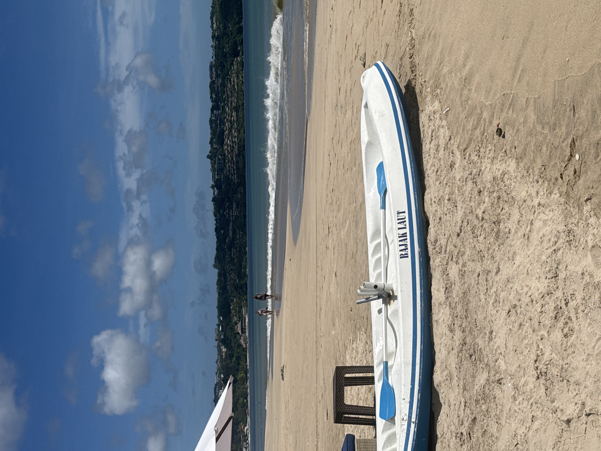 A kayak is lying on the sand at Pandawa Beach in Kuta Selatan, Indonesia. A couple is walking along the shoreline. The sky is blue with scattered clouds, and the ocean is visible. A small table and partial view of a white umbrella are also visible.