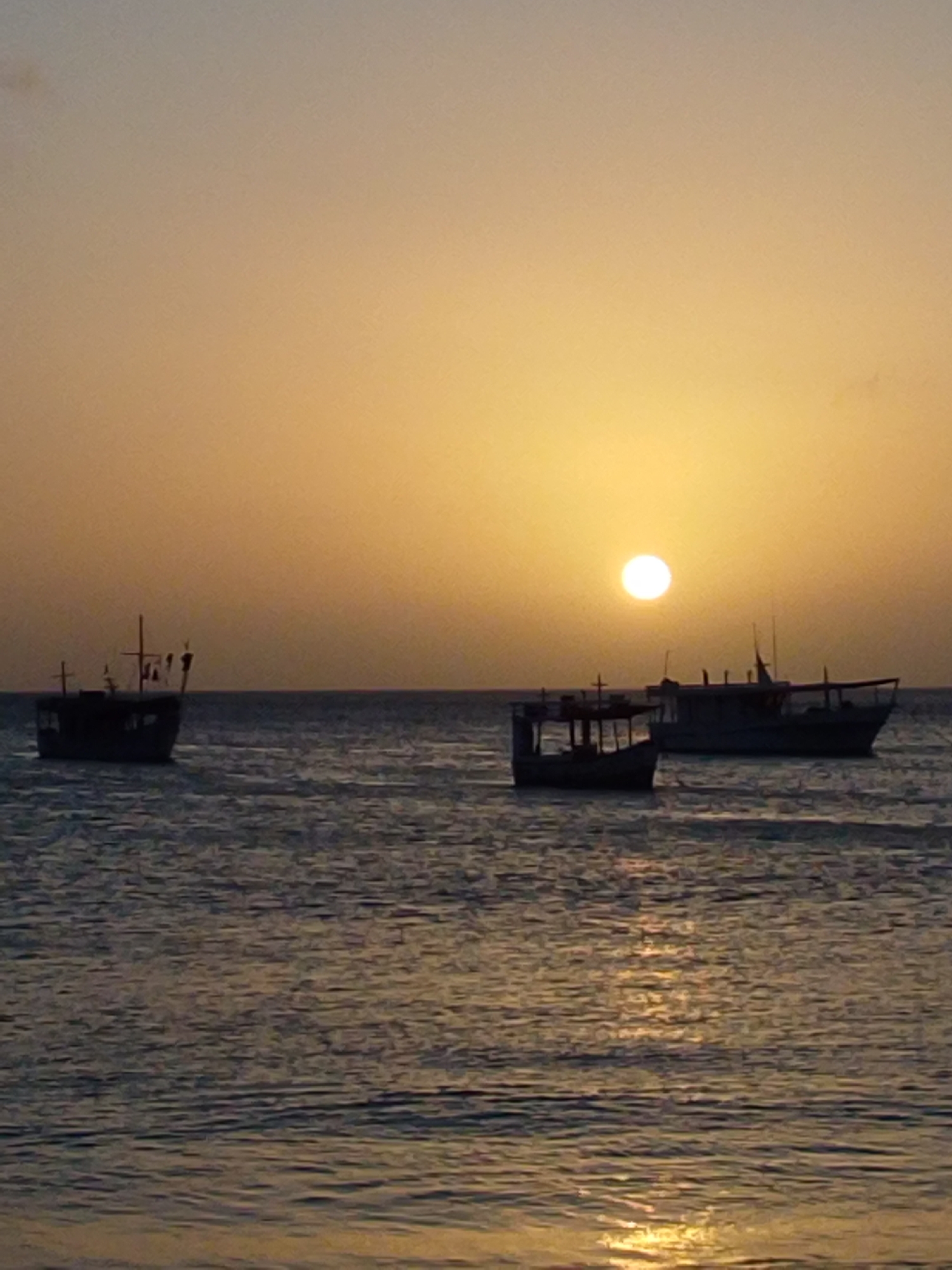 This is a photograph taken at dusk in the city of Juan Griego, Venezuela, featuring a tranquil ocean scene. The sun, a bright orange orb, is setting on the horizon, casting a warm, golden glow across the sky and reflecting on the gently rippling water. In the foreground, the sea exhibits a textured surface with shimmering highlights from the setting sun. Silhouetted against the vibrant sky are three fishing boats. The boat on the far left is smaller and has a simple, open structure with a mast. To its right, and slightly further back, is a larger boat, also silhouetted, with a more complex superstructure and a prominent mast. The third boat, positioned to the right of the larger vessel, is similar in size to the middle one and also appears to be a fishing boat. The overall atmosphere is serene and peaceful, capturing the essence of a quiet evening by the sea in this Venezuelan coastal town. There are no people visible in the photograph.