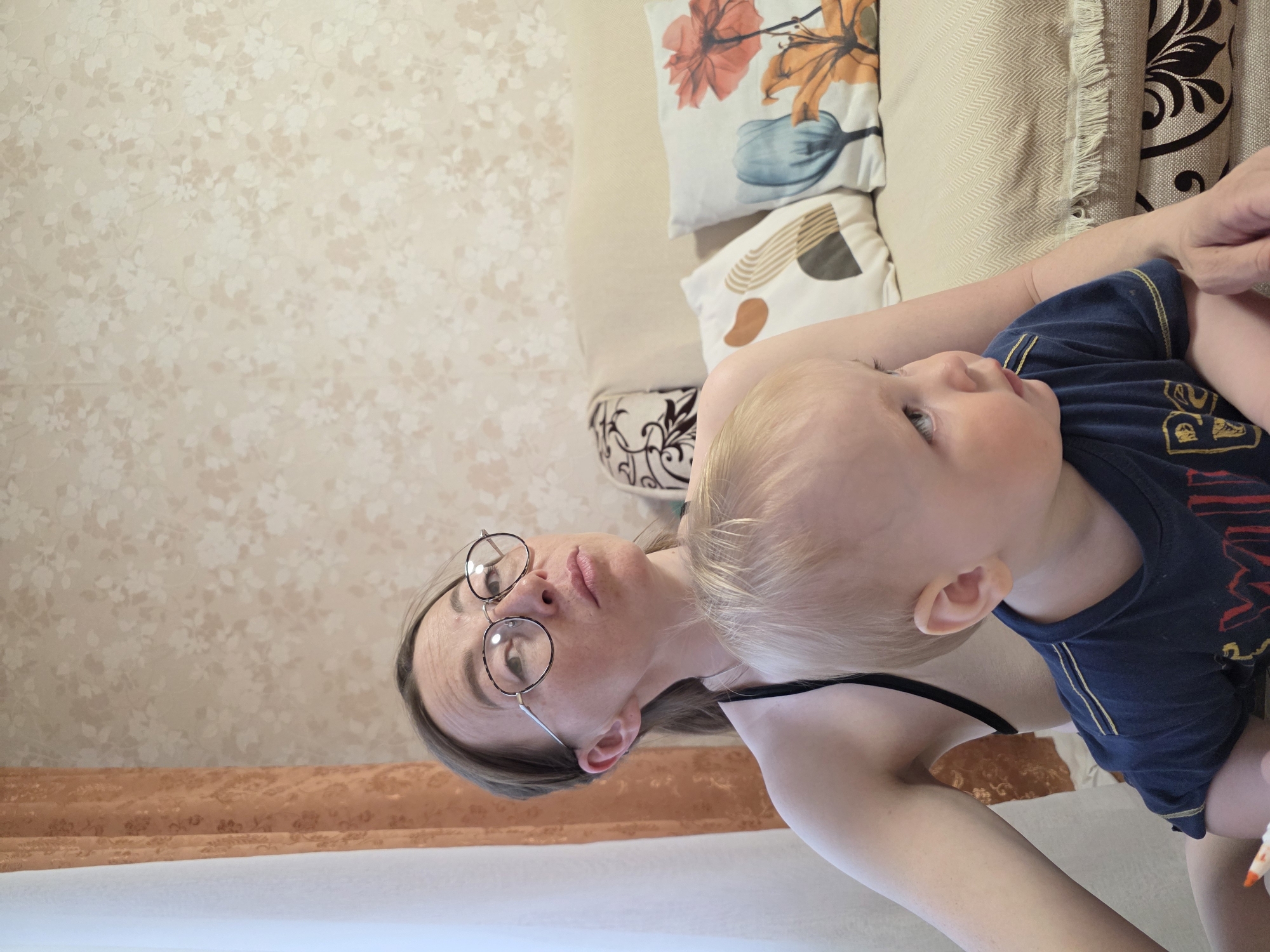 A fair-skinned woman with round glasses is holding a fair-skinned toddler with blond hair, both are indoors in what appears to be a residence, possibly located in Chaikovsky, Russia based on prior context. The woman is lying on what appears to be a couch with decorative pillows, and the toddler is in her arms, wearing a blue shirt. The background consists of a textured wallpaper. The scene seems candid, possibly a moment between mother and child.