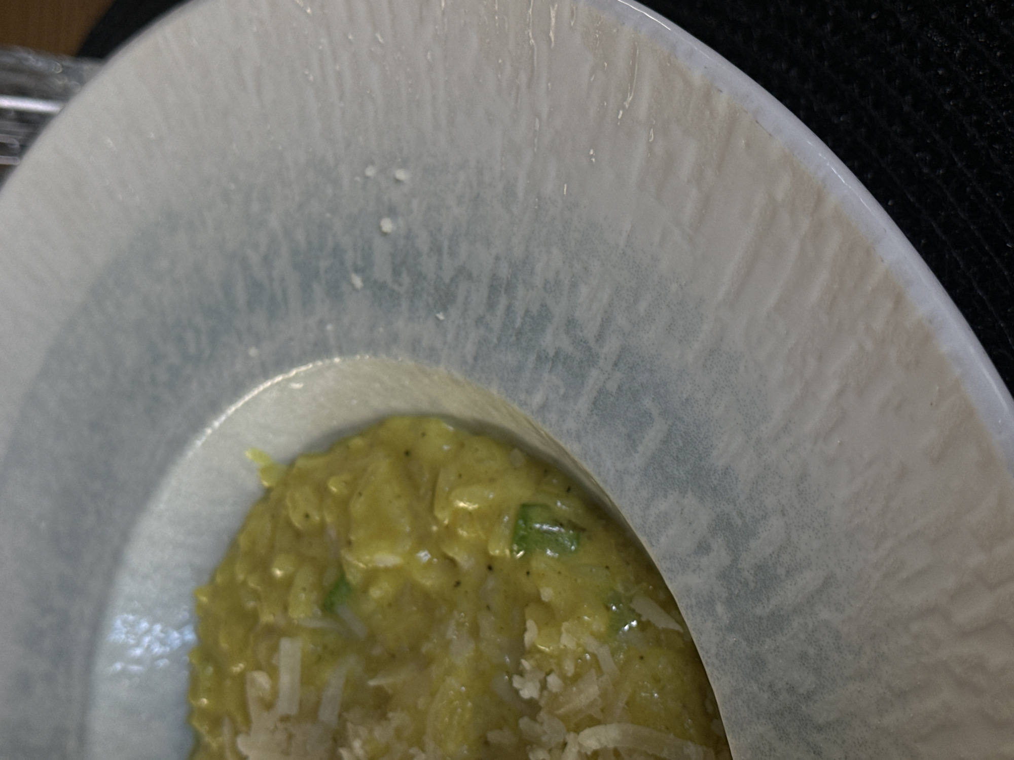 A close-up shot shows a portion of a pale blue and white speckled plate. Visible within the plate is a serving of yellow risotto with scattered white grated cheese. The edge of a black textured surface is visible near the top right of the frame. The location cannot be determined from the image, and there is no information related to people or interactions.