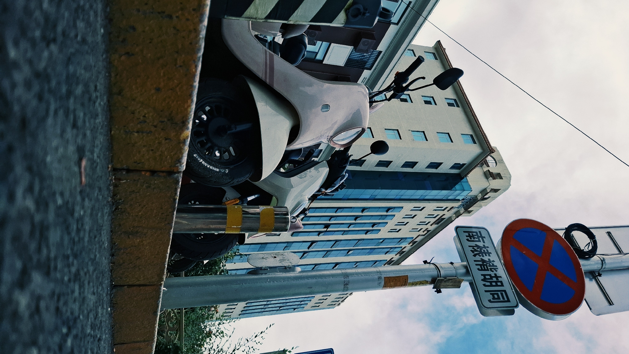 A street scene in Dongcheng District, China, is depicted from a low angle. A modern, white-and-beige scooter is parked parallel to a curb with a yellow line. A high-rise building with reflective blue glass windows rises in the background. A street sign displaying a "no parking" symbol in blue and red is visible. A sign with Chinese characters reading "南麓禧胡同" (Nánlù xǐ hútòng) is attached to the same pole. The sky above is partly cloudy.