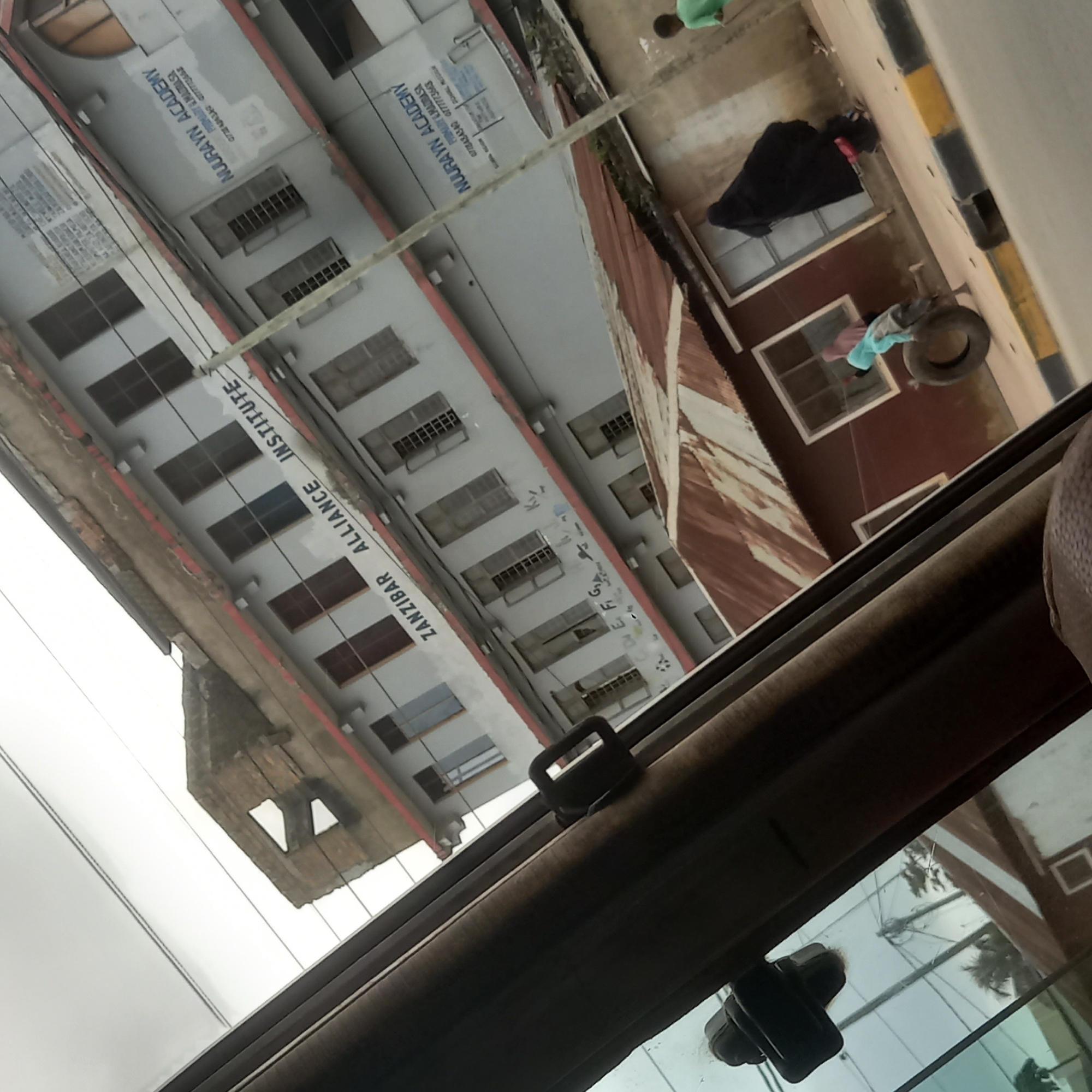 The image shows a street scene in Bububu, Tanzania, with two buildings visible. The foreground is dominated by the interior of a vehicle, with a dark brown curtain and a black seatbelt buckle visible. The exterior view through the window reveals two buildings. The larger building, on the left, is a multi-story structure with light grey walls and red trim. It appears to be an educational institution, with signage that reads "ZANZIBAR ALLIANCE INSTITUTE" and "NUURAYN ACADEMY". The building has numerous windows, some with shutters, and visible wiring. A partially demolished section is also apparent at the top left of this building. To the right of this building, a smaller, single-story structure with a rusty corrugated metal roof is visible. In front of this smaller building, there are at least three people. One person is wearing a black abaya, and another, a child wearing a light blue garment, is interacting with a large tire on the sidewalk. Another child, wearing a green shirt, is visible further down the street. Electrical wires are strung across the scene, and a street curb with yellow and black markings is present in the lower right. The sky is overcast and white.