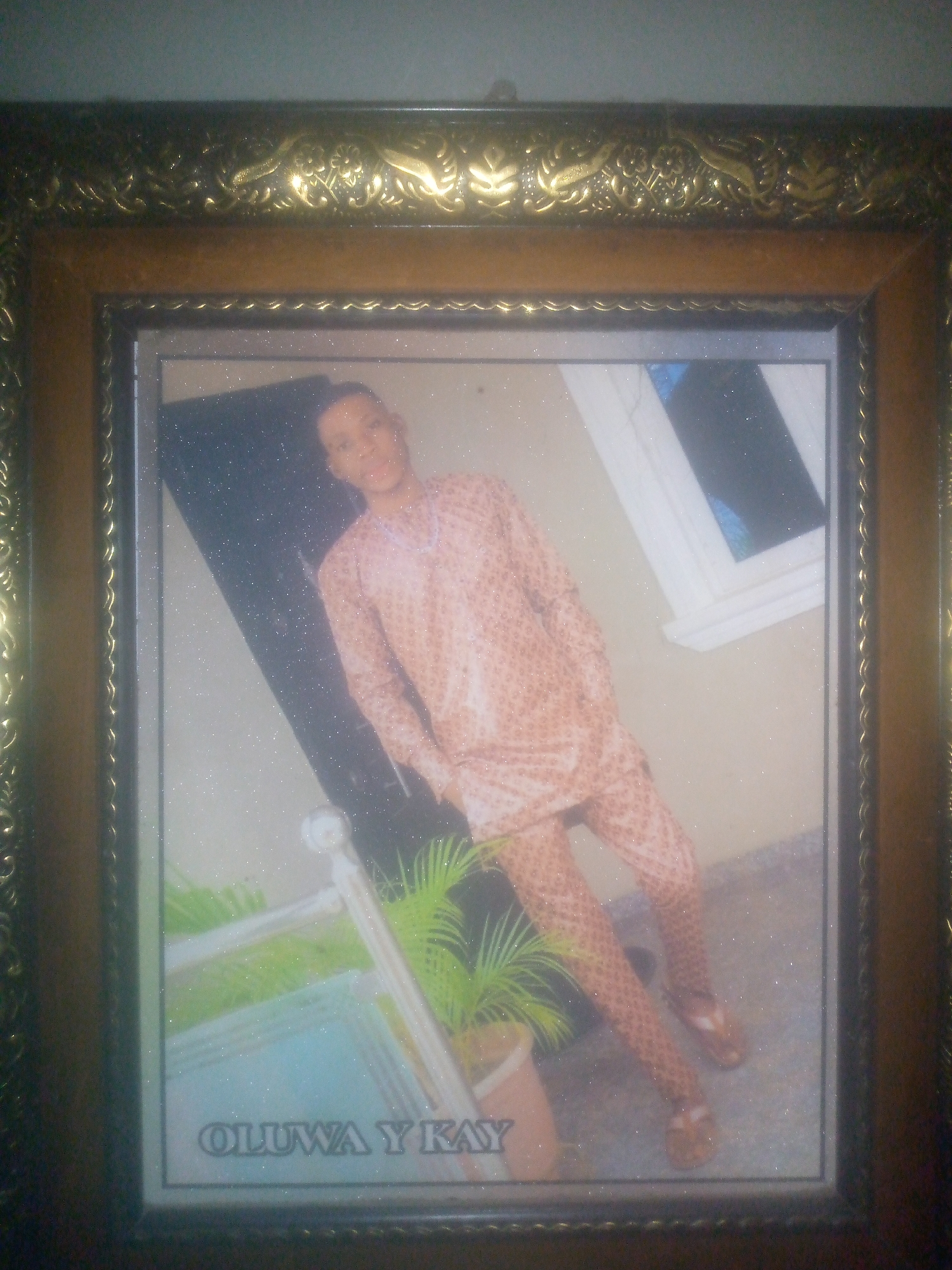 A framed photo of a person standing in front of a dark background and window. The person is wearing a patterned shirt and pants. There is a potted plant in the lower left corner of the frame. The name "OLUWA Y KAY" is printed at the bottom of the photo. The frame is ornate and gold in color. The city and location are unknown.