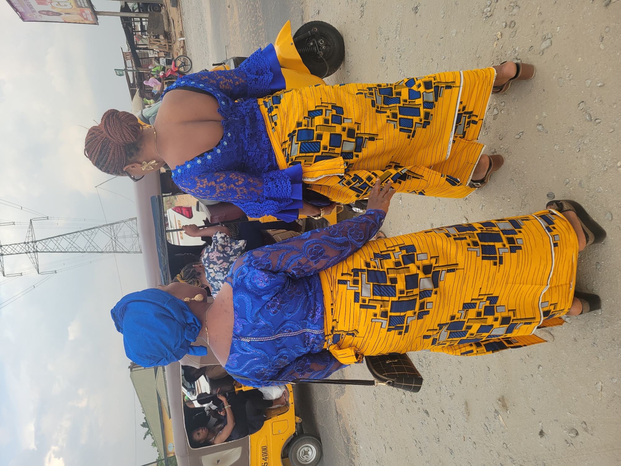 Two women in matching blue lace tops and yellow and blue patterned skirts stand on a cracked concrete surface near a yellow commercial tricycle (a "keke") in Kwamba, Nigeria. The women are viewed from behind; one has her hair braided, and the other wears a blue headwrap. The tricycle appears to be transporting passengers. Overhead power lines are visible in the background, along with other buildings and street scenes.