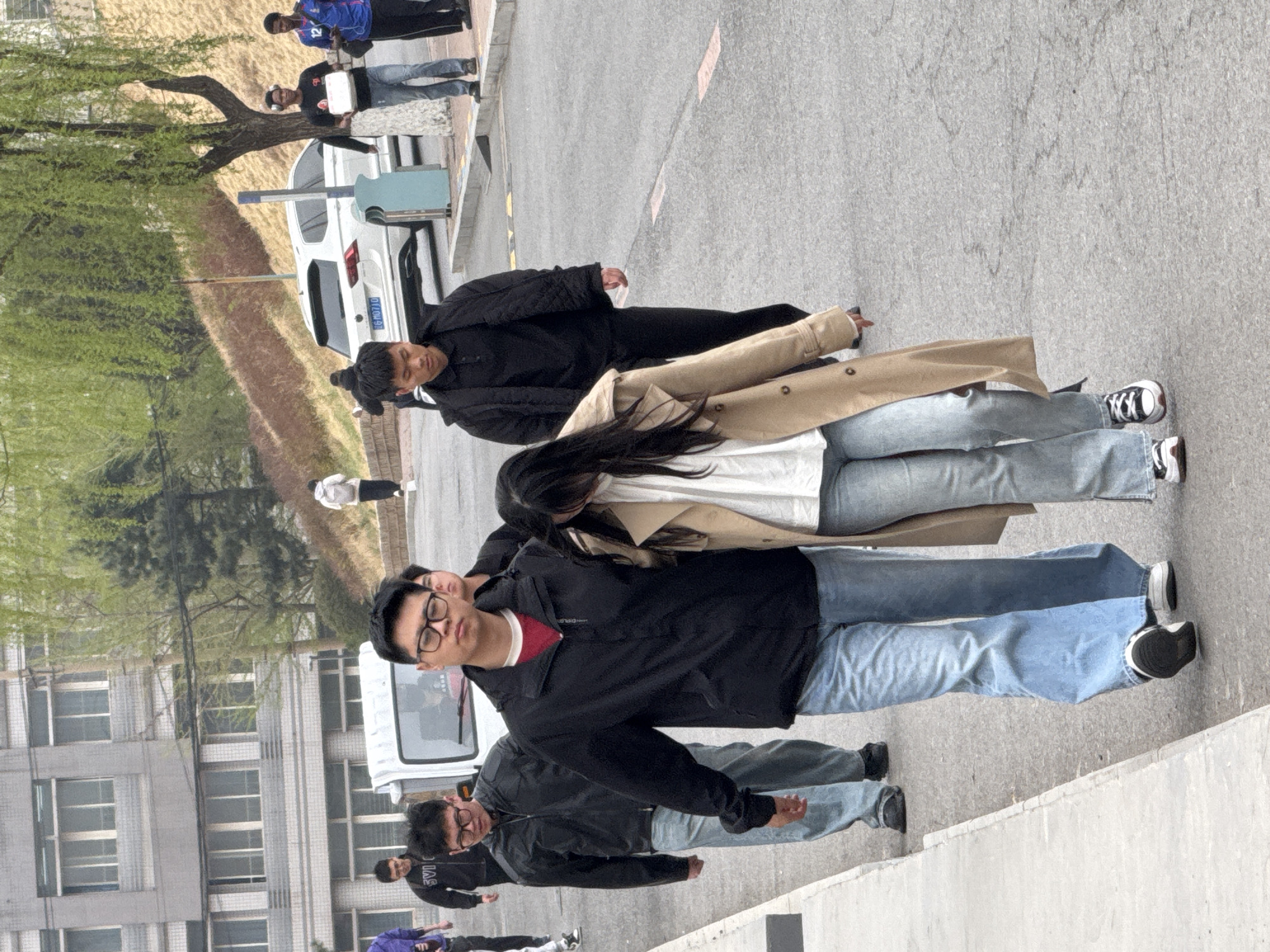 The image shows a group of people standing on a paved surface in Zhongtun, China. A man wearing glasses and a black jacket over a red shirt is at the forefront. Behind him are more people in similar jackets and jeans. A woman in a tan trench coat stands behind him. A white van is parked nearby, and other vehicles are visible in the background. A building with multiple windows is seen on the left. A grassy area with trees is on the opposite side of the road. The scene appears to be a casual gathering on a street in a city.