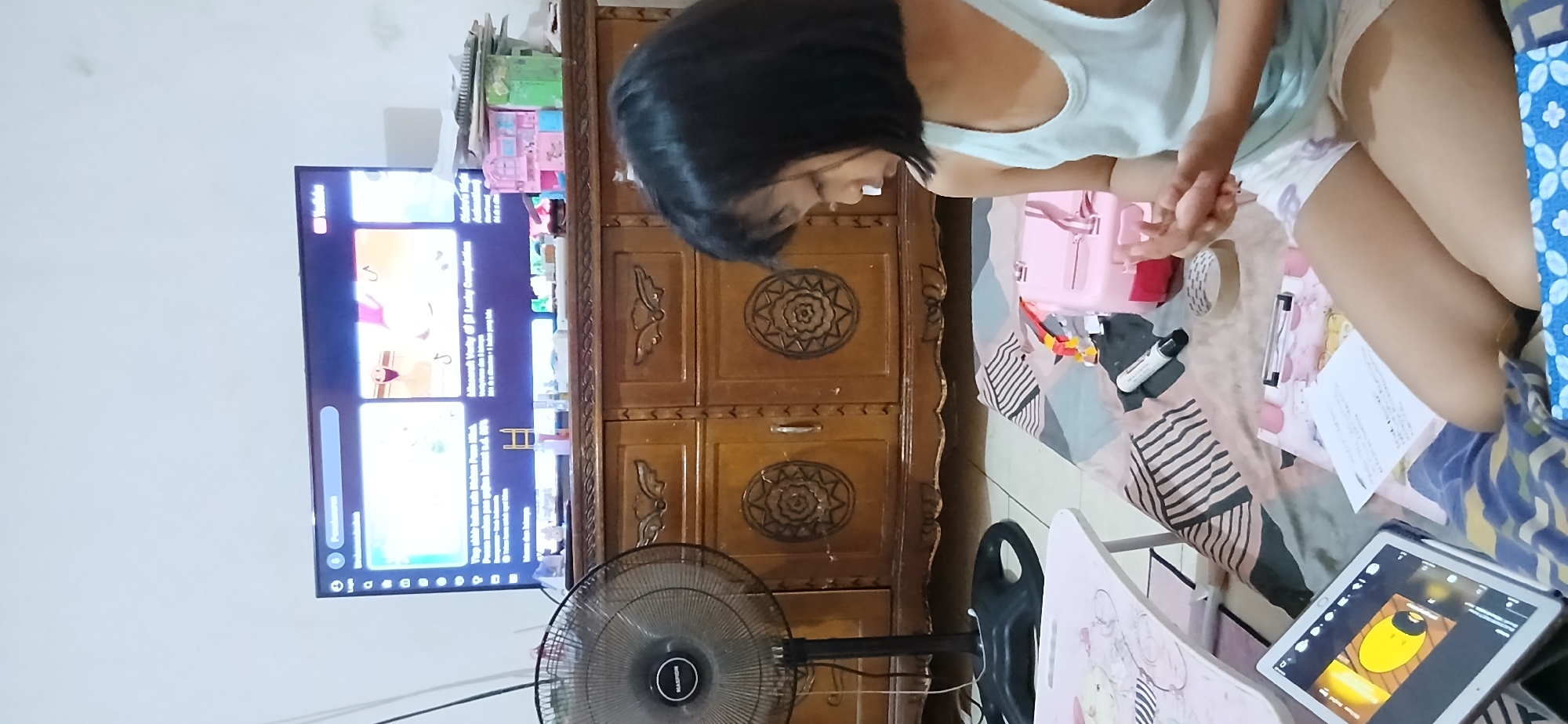 The image captures an indoor scene, likely a living room or bedroom in Samarinda, Indonesia, given the context provided. In the foreground, a young girl with short black hair is sitting on a patterned bedspread, facing to her right and looking down at her hands. She is wearing a light blue tank top and white shorts with a subtle pattern. Her hands are clasped together.
To the left of the girl, a white tablet displays a cartoon image. In front of the girl are various items: a pink toy box, a roll of tape, a black marker, and a clipboard holding some papers. Behind her is a dark wooden cabinet with ornate circular carvings on the doors. To the left of the cabinet is a black oscillating fan. Above the cabinet, a large flat-screen television displays what appears to be a YouTube interface with video thumbnails. The overall environment suggests a casual, domestic setting.
