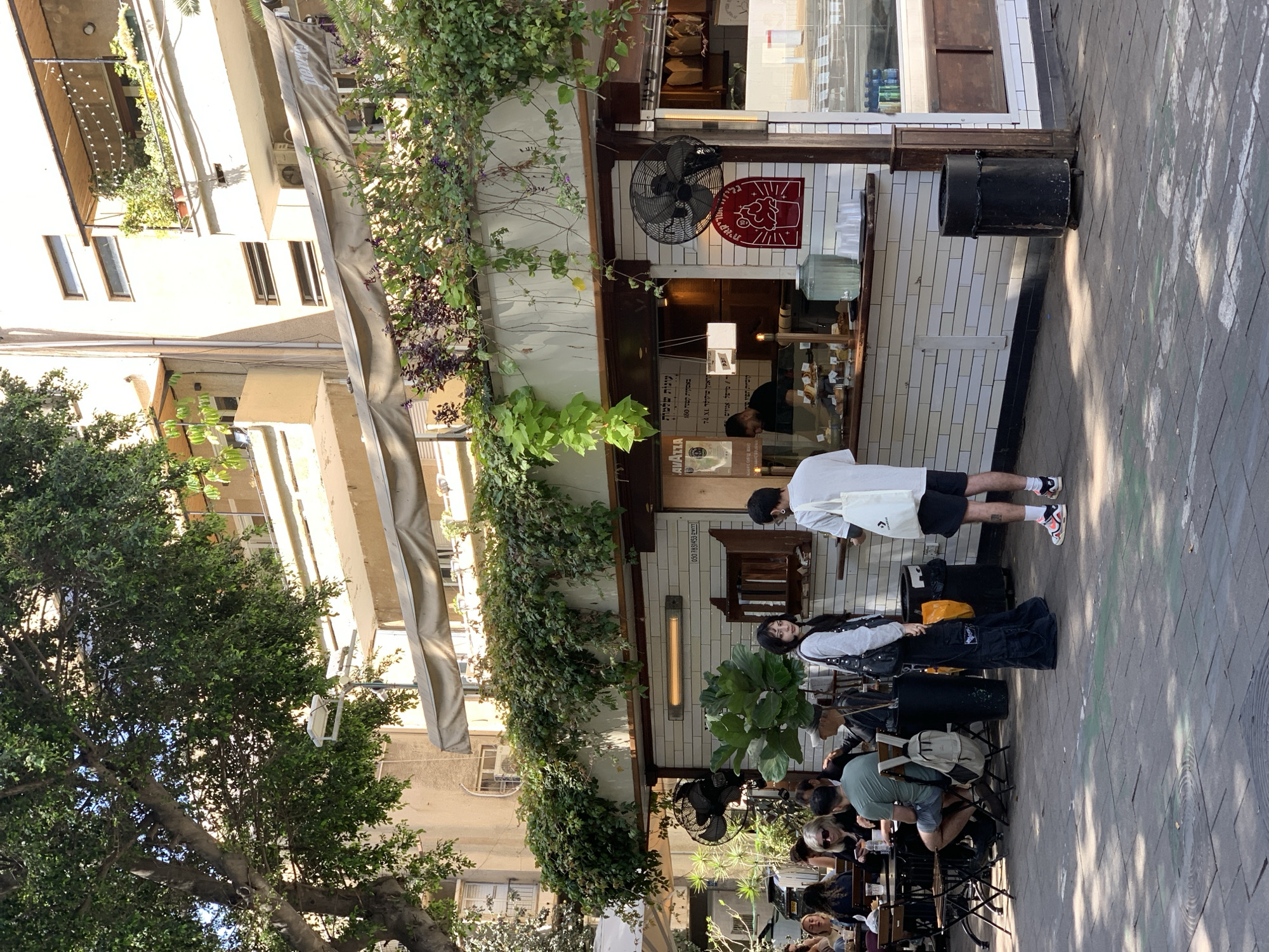 The image depicts a street scene in Tel Aviv, Israel, featuring an outdoor cafe. Several people are seated at tables, some are dining. A man in a white shirt and black shorts is standing at the cafe counter, possibly ordering. Another person in a grey top and black cargo pants appears to be waiting. The cafe facade is decorated with greenery and has an awning providing shade. A fan is mounted on the wall outside the cafe. There is a red and white logo of a coffee mug and face. The building behind the cafe appears to be multi-story, with windows and architectural details. A tree with dense foliage is on the left side of the frame.