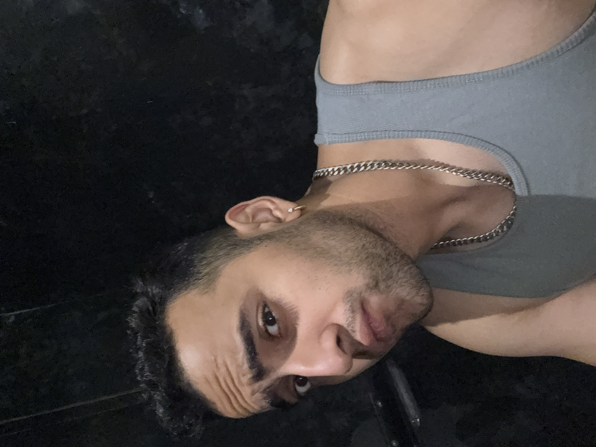 This media file features a close-up portrait of a young man, presented in a rotated orientation, making him appear on his side. The subject is a man with dark hair, short stubble or a beard, and dark, prominent eyebrows. He is wearing a grey sleeveless tank top and a silver chain necklace. A small gold hoop earring is visible in his left ear.
The man's gaze is directed towards the viewer, and his expression appears serious or intense, with a slight furrow in his brow. His left eye is clearly visible and open, while his right eye is partially obscured but also appears open.
The background is uniformly dark and appears to be black, providing no specific details or elements. This dark background suggests either a nighttime setting or a dimly lit indoor environment. The lighting on the man's face and upper body seems to be artificial, casting some soft shadows. No specific location cues are visible within the image itself to confirm the Kolkata, India context. There is no visible text. The overall atmosphere of the photo is intimate and focused on the subject.