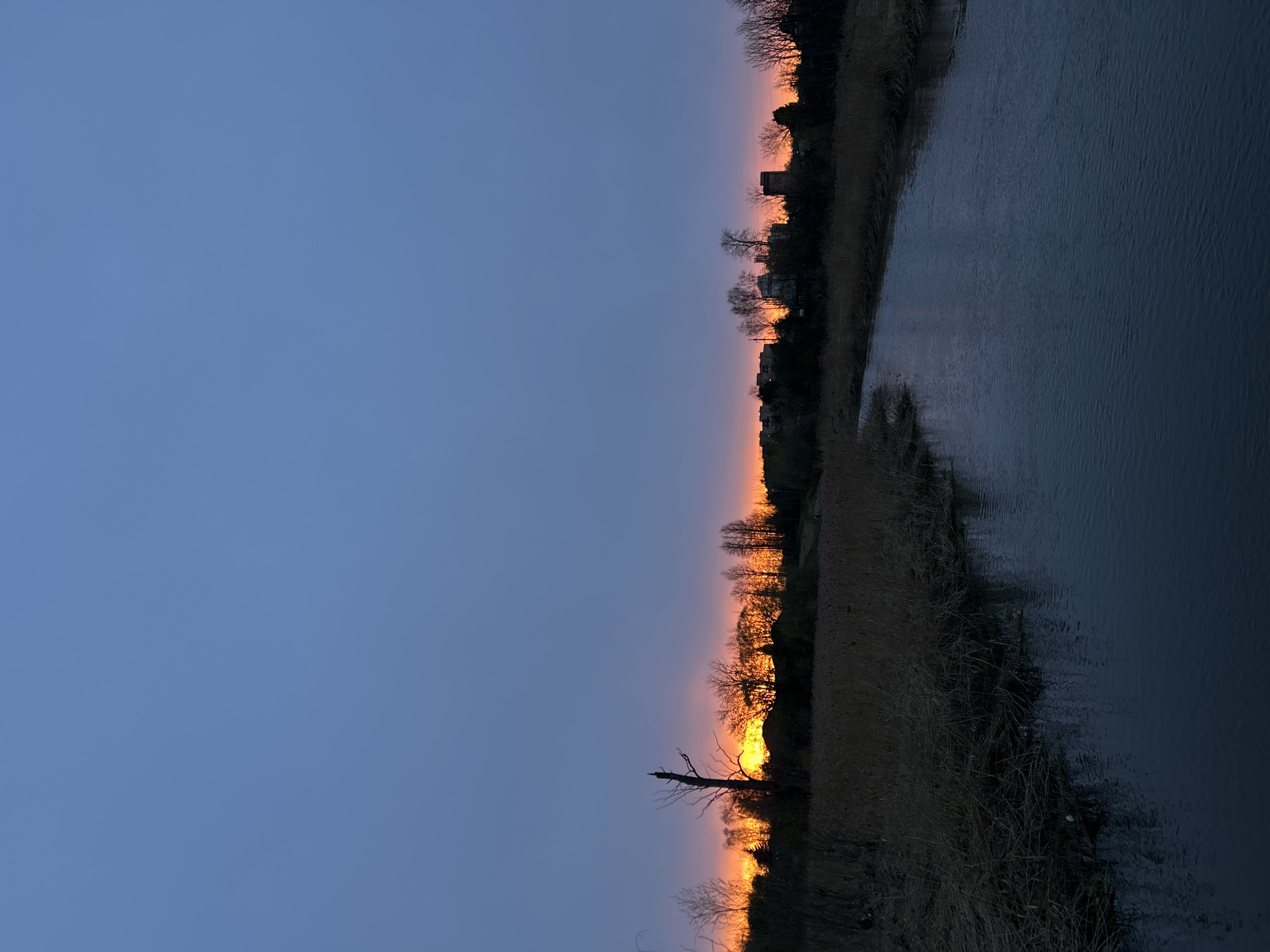 The image depicts a landscape scene at twilight or dusk, possibly in Klin, Russia. The right side of the image shows water, likely a river or lake, with gentle ripples on its surface. The opposite bank features a strip of vegetation and trees silhouetted against the horizon. A band of orange and yellow hues paints the sky above the treeline, suggesting a sunset or sunrise. The sky transitions to a deep blue color above the horizon. In the foreground, there's a leafless tree trunk standing prominently against the colorful sky. The photograph has a vertically oriented composition, with the sky taking up the majority of the frame. The location is assumed to be Klin, Russia based on provided details.